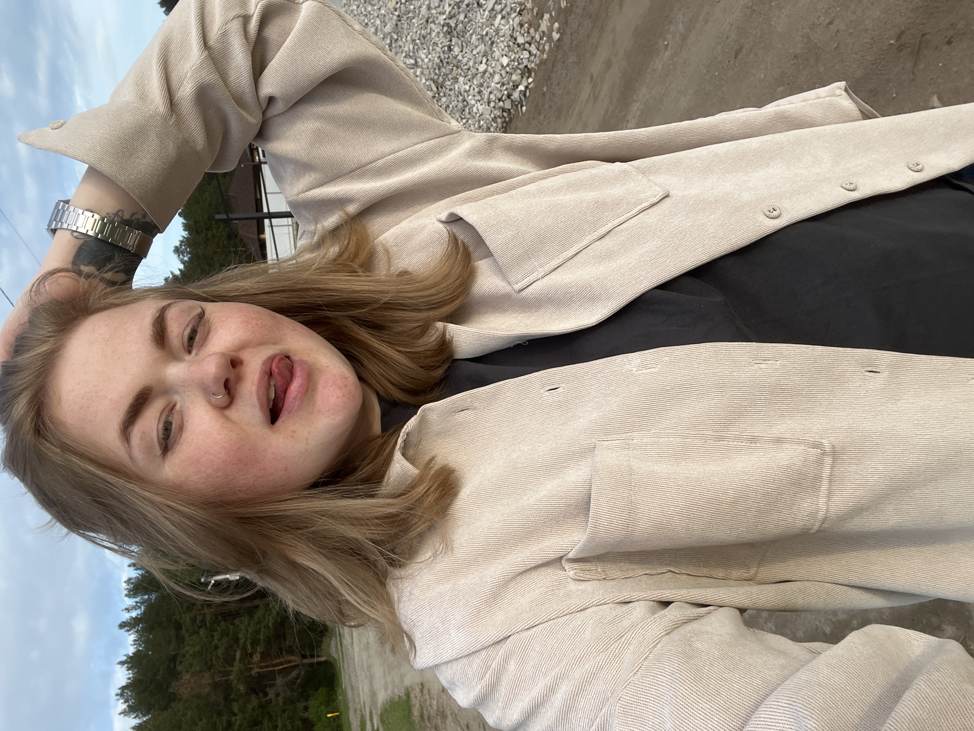 A young woman with blonde hair, freckles, and a nose ring is posing for a selfie outdoors. She has one hand on her head and is sticking her tongue out. She is wearing a black shirt and a beige corduroy jacket. A silver watch and a tattoo are visible on her left wrist. In the background, there are trees, a building, gravel, and part of a road. The location is Chaikovsky, Russia, based on the landscape.