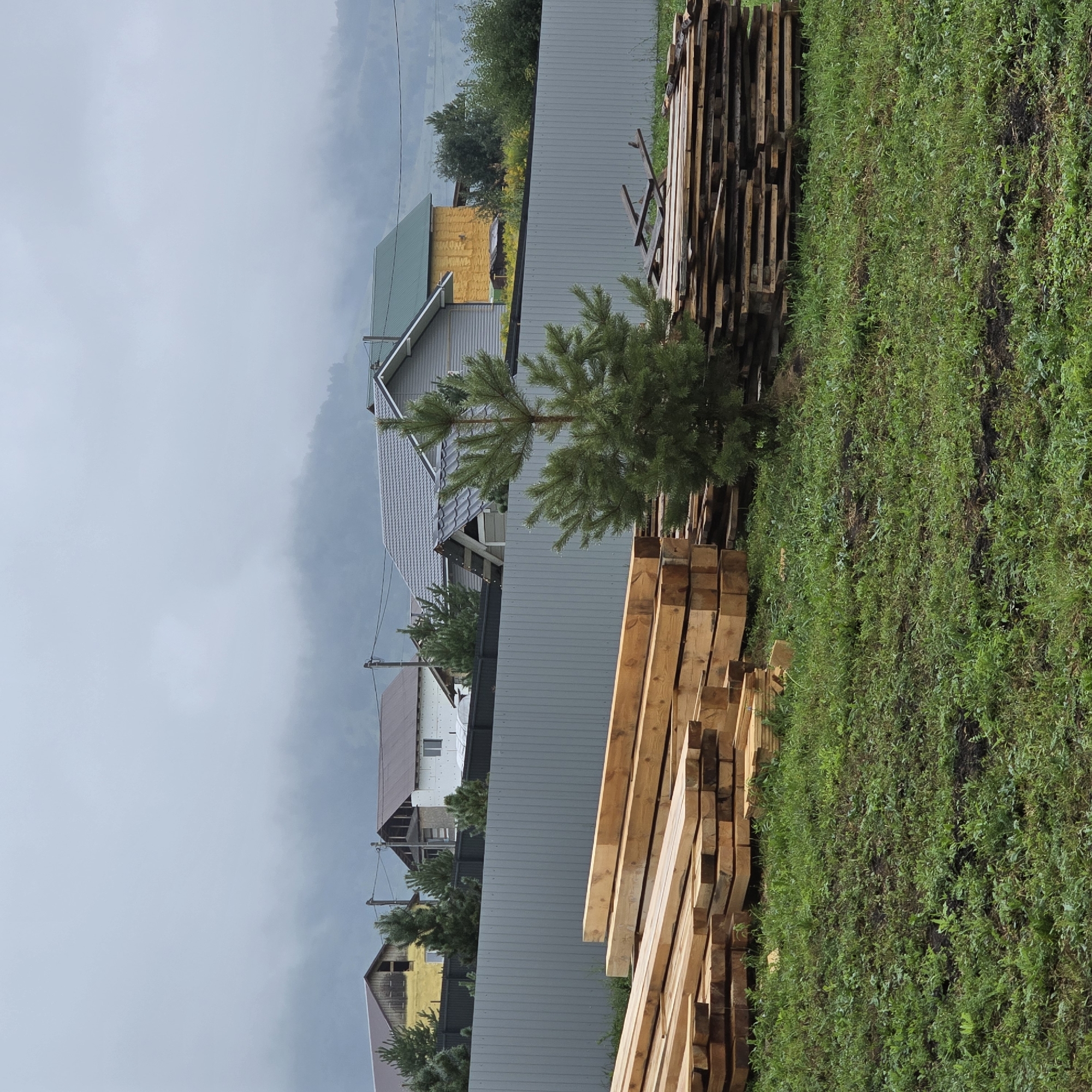 The image depicts an outdoor scene in Belokurikha, Russia, on a cloudy day. In the foreground, piles of lumber are stacked on a grassy area. To the left, a large stack of squared timber beams is prominent, while to the right, thinner planks are stacked. A small pine tree stands between the stacks of lumber. Behind the lumber and tree, a tall, light gray metal fence runs horizontally across the middle of the image. Beyond the fence, several houses are visible. The houses have various roof colors, including brown, gray, and green, and some are under construction with exposed framing and insulation. The background features a misty, mountainous landscape shrouded in fog. Power lines are visible running between utility poles. The overall impression is one of rural construction or a staging area for building materials in a mountainous, possibly remote, location. There are no people or animals present in the image.