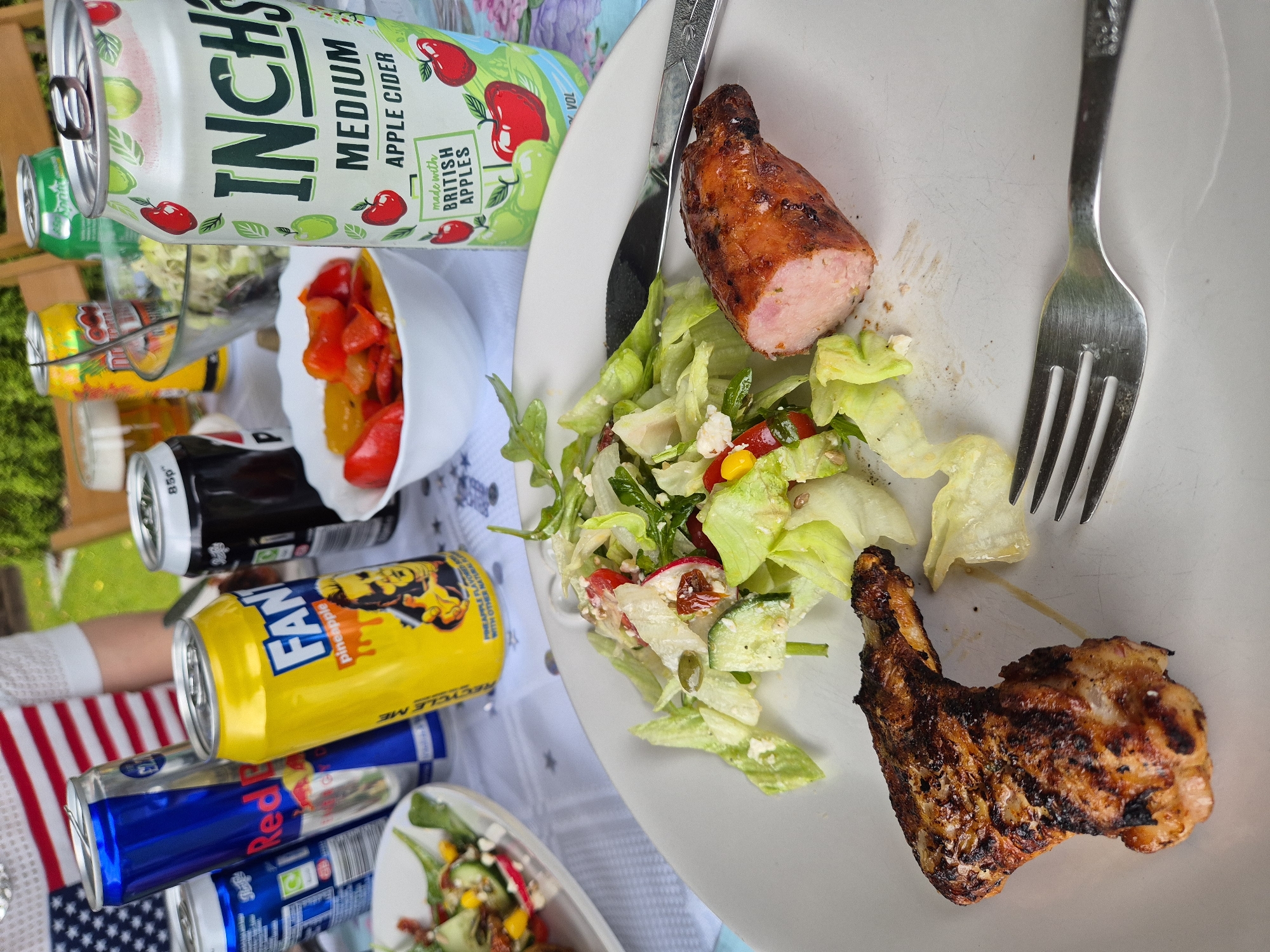 Here's a description of the image:
**Scene:** The image shows a partly eaten plate of food on a table. The food includes a sausage cut in half, a piece of grilled chicken, and a mixed salad with lettuce, radishes, corn, and other ingredients.
**Objects:**
* A white plate with food
* Fork and knife
* Cans of various drinks: Inch's cider, Fanta Pineapple, Red Bull, and others that are partially visible.
* A bowl of roasted peppers.
* A table with a tablecloth with stars on it.
**Subjects:** The focus is on the partially eaten meal. A person's arm wearing a white shirt with a part of the American flag is partially visible in the background.
**Environment:** The scene appears to be outdoors, possibly a picnic or a barbecue setting.
**Location:** The presence of Inch's Cider indicates the West Suffolk, United Kingdom.
