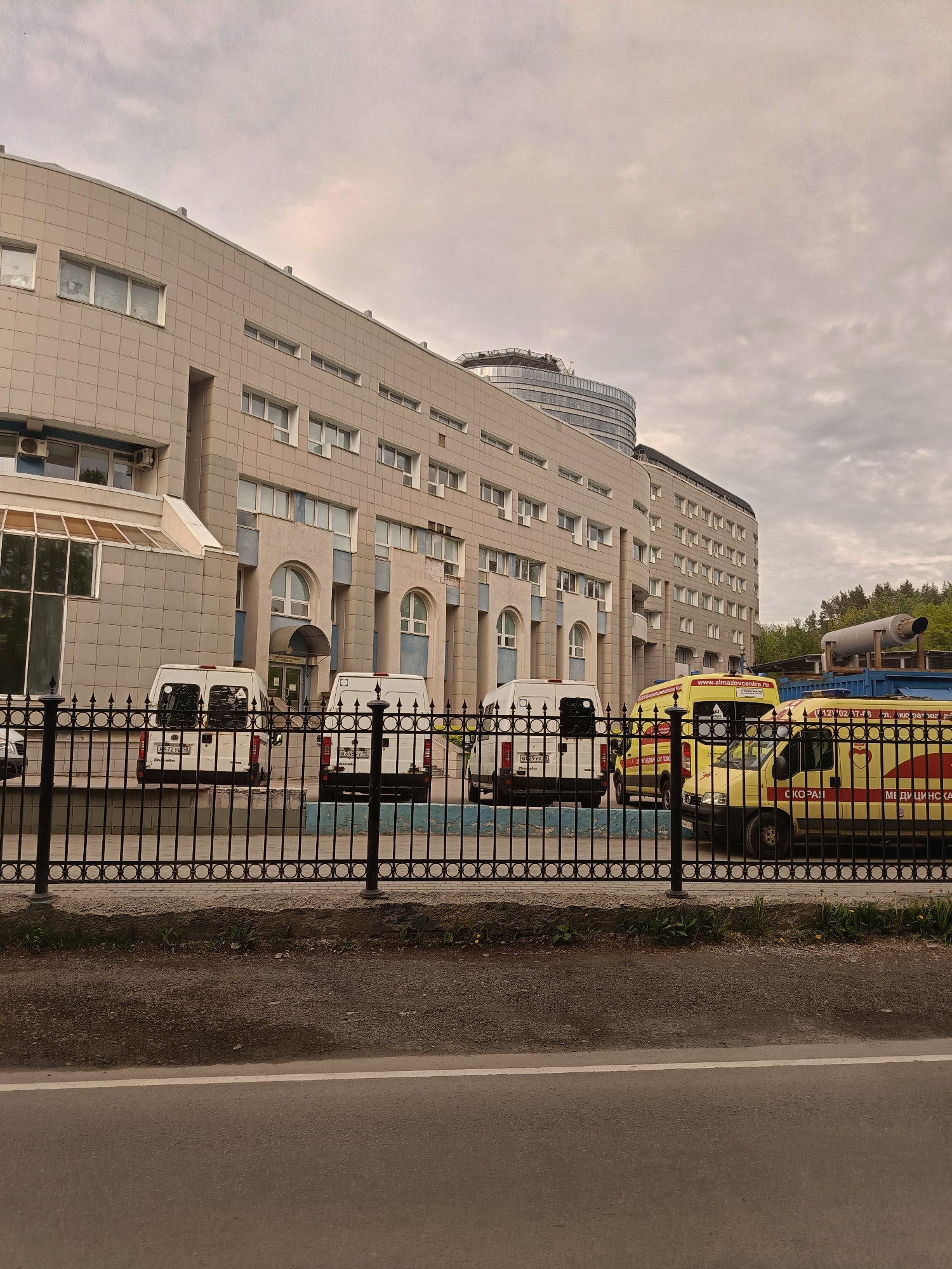 The image depicts the Almazov National Medical Research Centre in Saint Petersburg, Russia. In the foreground, a black metal fence separates the viewer from the building and vehicles. Behind the fence, several vehicles are parked, including white vans and yellow ambulances. The ambulances have Russian text on them, including "СКОРАЯ МЕДИЦИНСКАЯ ПОМОЩЬ," which translates to "Emergency Medical Service." The Almazov Centre building is a large, multi-story structure with a beige facade and many windows. A cylindrical building is visible behind the main structure, possibly a newer addition. The sky is overcast, and a road with a white line is visible in the extreme foreground. The scene appears to be taken from across the street from the medical center.