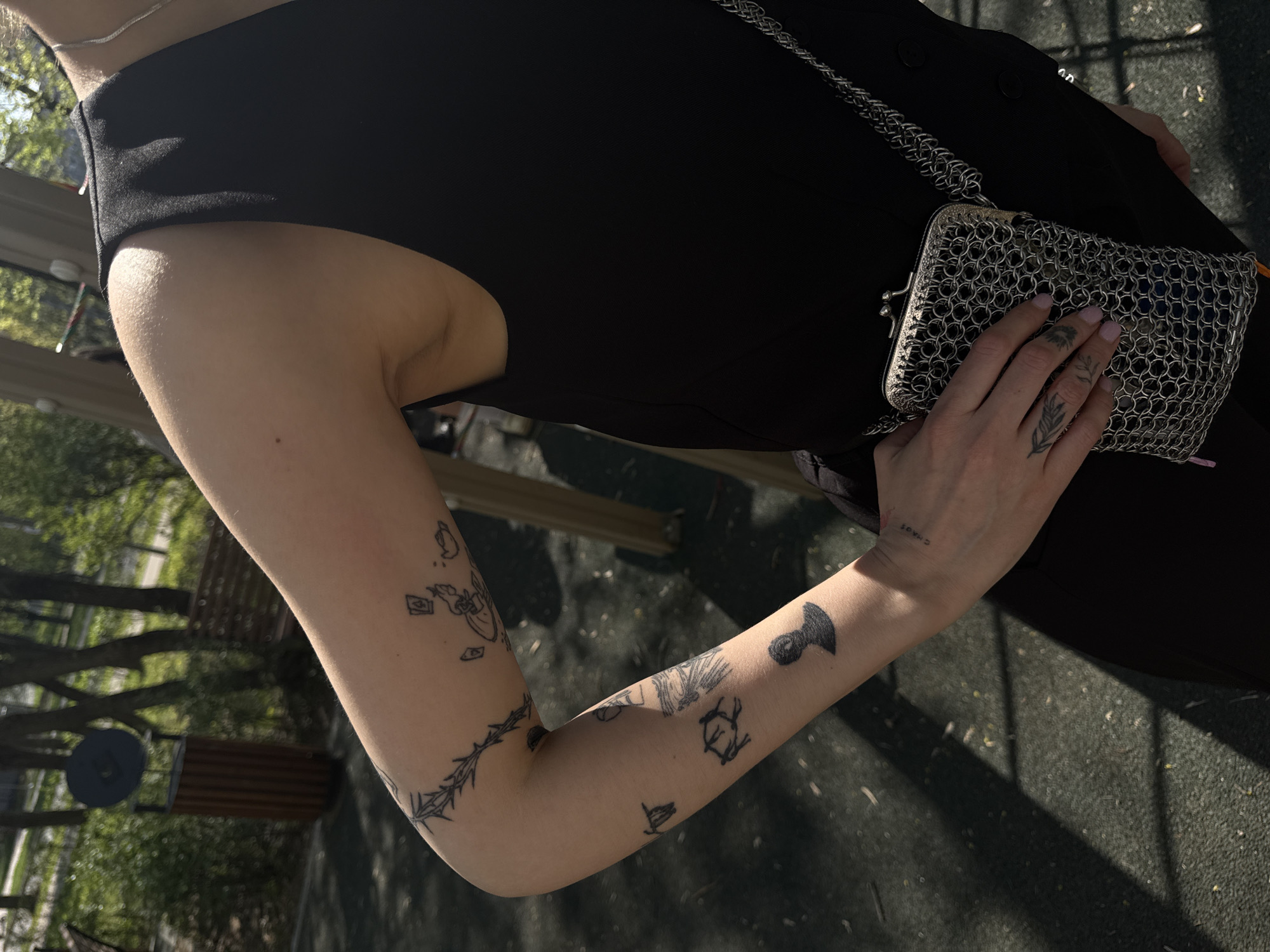 A woman is standing outdoors, wearing a black sleeveless garment. She is holding a silver metal mesh bag with a chain strap. Her arm is visible and has several tattoos on it. The background appears to be a park or outdoor recreational area. The image may be taken in the city of Moscow, Russia due to the presence of park benches and tree varieties common in the region.