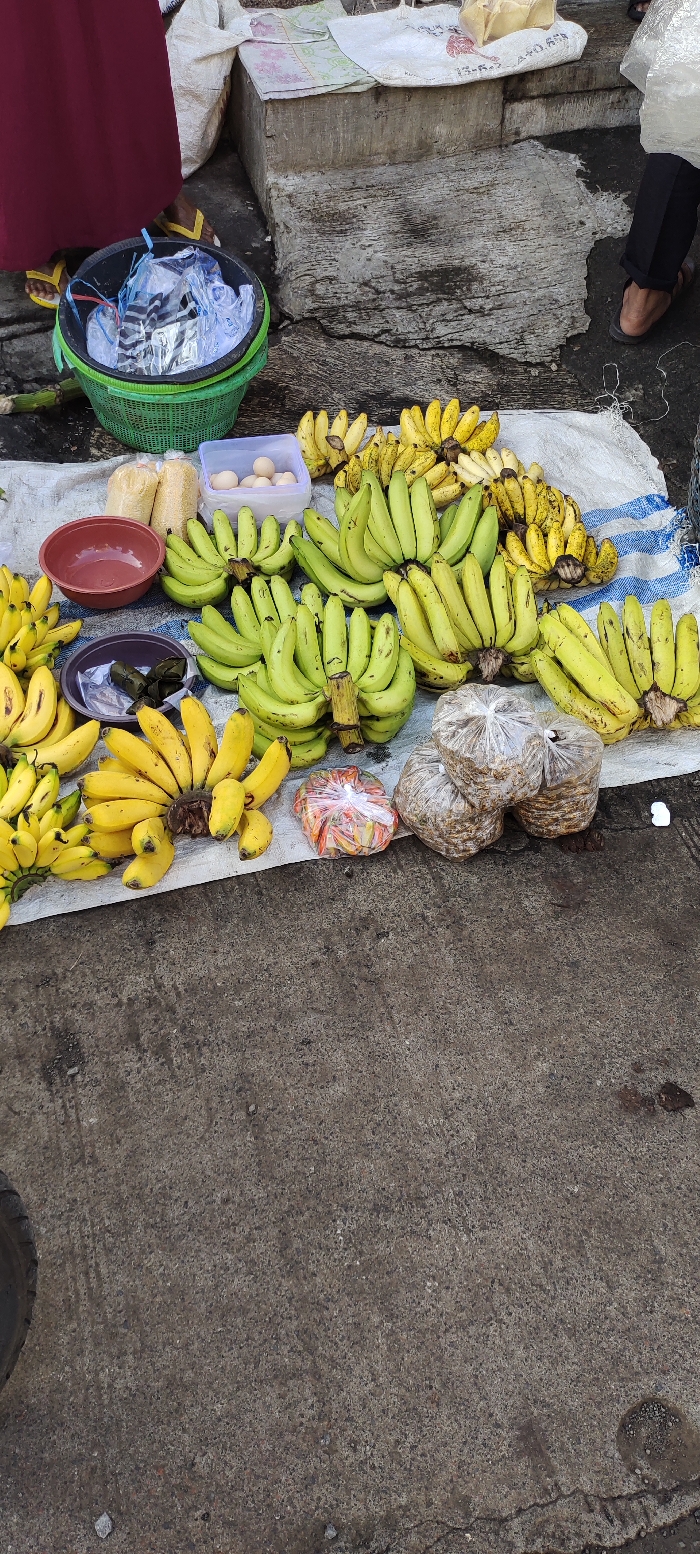 The image shows a street vendor selling bananas and other snacks in Kecamatan Lawang, Indonesia, at the coordinates -7.846 latitude and 112.7 longitude. The vendor has laid out a large white cloth on the ground, on which they have arranged bananas, eggs, snacks wrapped in plastic, and a few bowls. The bananas are arranged in groups, some ripe and yellow, some still green. The street is paved with gray concrete, with a few cracks and some small, dark patches on the surface. A partially visible tire sits on the left side of the image, with a blurry figure in the upper left. An individual's feet are visible on the right side of the image. The scene appears to be a typical street market setting in Indonesia.