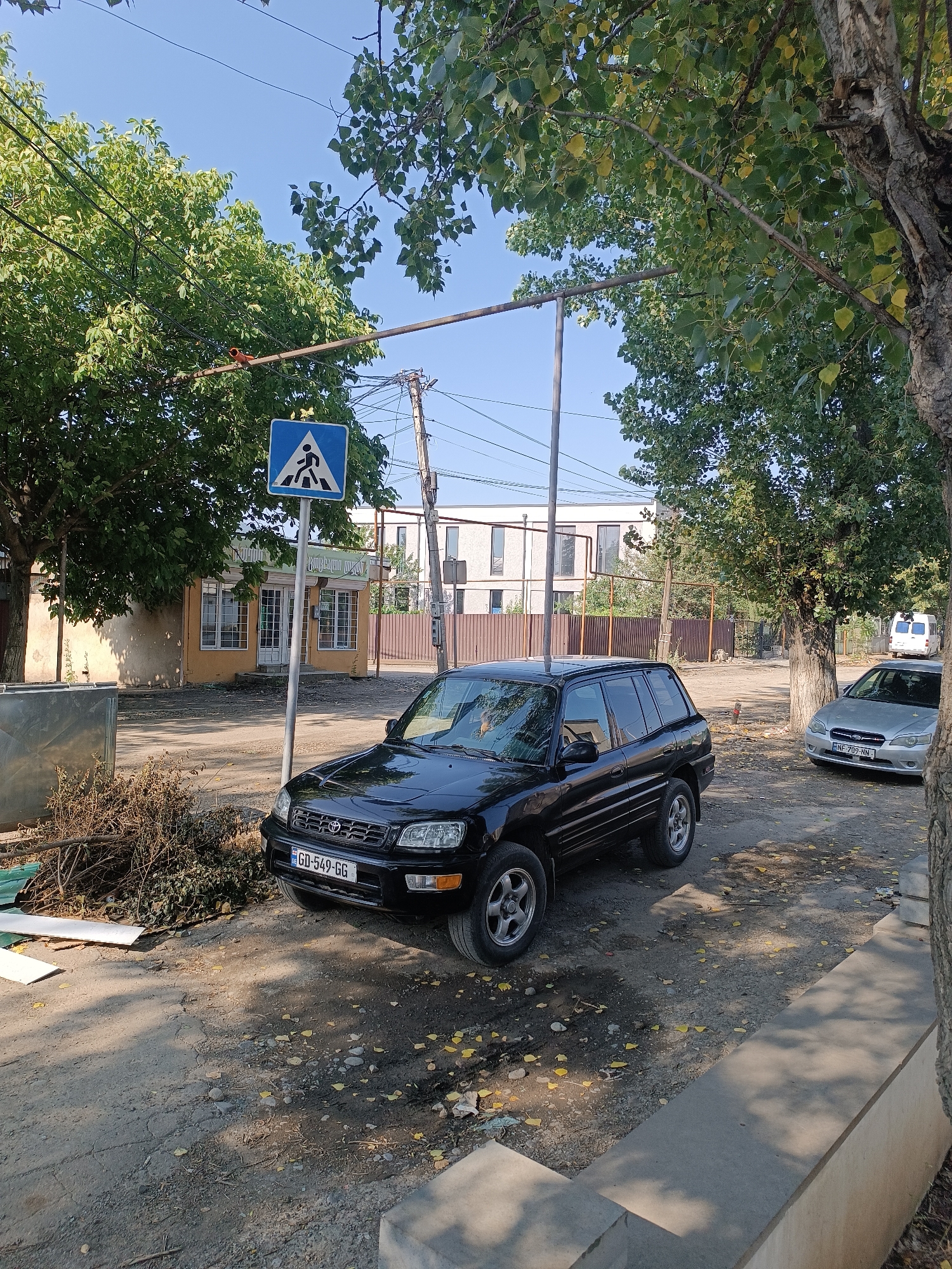 In Khashuri, Georgia, a black Toyota RAV4 is parked on a dirt road lined with trees. A pedestrian crossing sign stands on the left side of the road, near a small yellow building. A silver Subaru Legacy is parked further down the road, and a white van is visible in the background. The sky is clear and blue, and sunlight filters through the leaves of the trees, casting shadows on the ground. There are scattered leaves on the road, suggesting it might be autumn. The scene appears to be a quiet residential street.