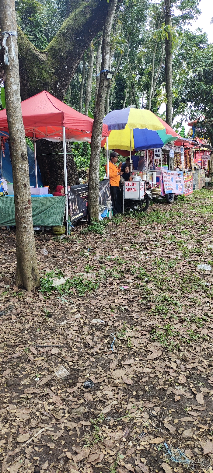 The scene is a street food vendor in Kecamatan Lawang, Indonesia, located at latitude -7.844 and longitude 112.688. There are two food stands side by side. The first stand has a colorful umbrella canopy and sells a variety of goods, including snacks and drinks. The second stand has a red canopy and appears to sell a variety of items, but the specifics are obscured by the camera angle. Three people are present: a man standing behind the first stand, a man standing in front of the first stand, and a woman standing behind the first stand. The ground is covered in fallen leaves. The stands are located in front of a large tree. There are several other trees visible in the background.