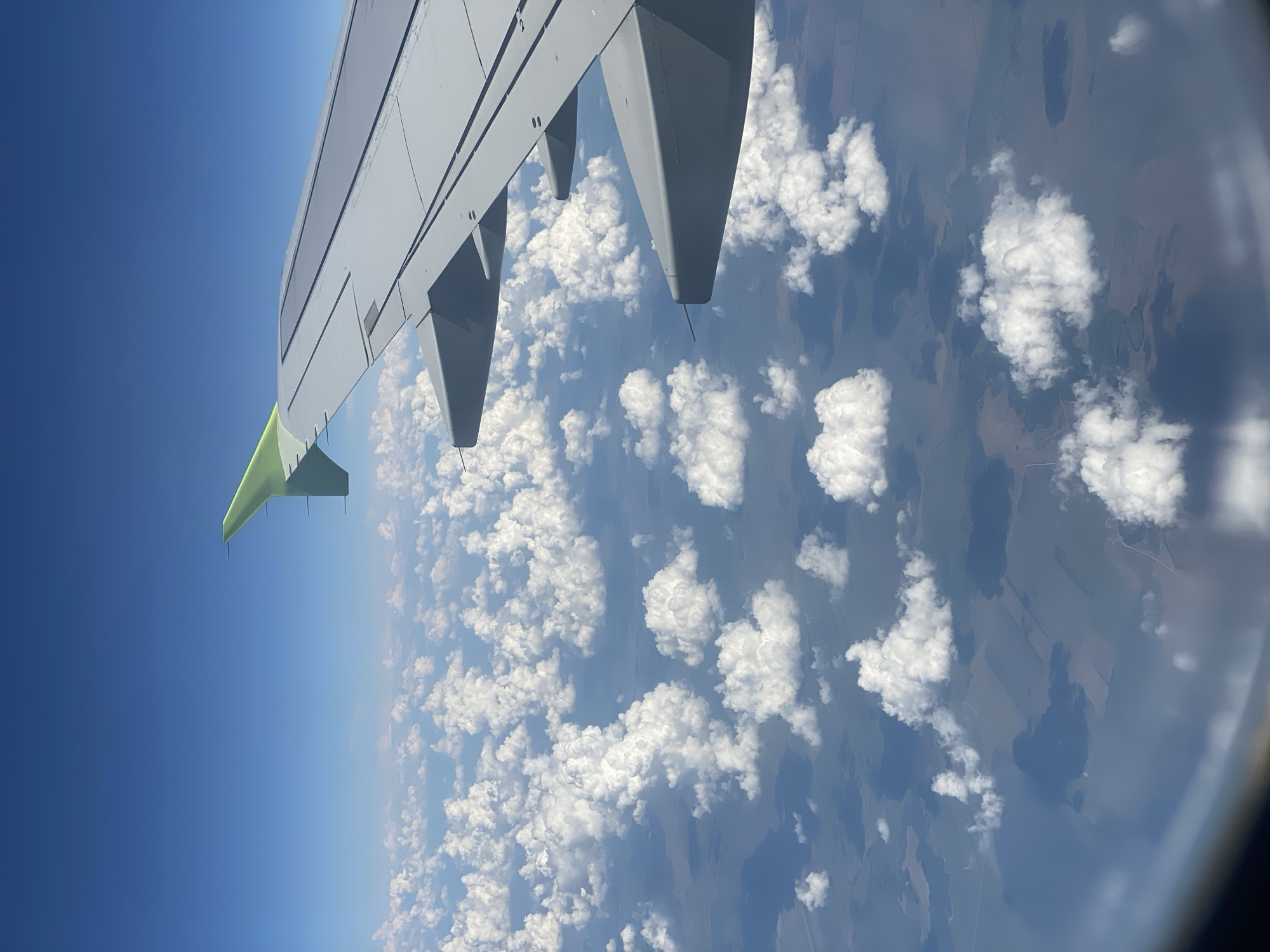 The image is taken from the perspective of an airplane window, looking down at a landscape below. The main subjects are the airplane wing, which occupies the left side of the frame and extends into the upper portion, and a vast expanse of cumulus clouds scattered across a visible ground. The ground appears to be a patchwork of fields and bodies of water, with some darker areas that could be forests or shadows.
The scene suggests the airplane is in flight, likely during the daytime due to the bright blue sky and the way the clouds are illuminated. The lighting indicates it's probably late morning or early afternoon. The weather appears clear with scattered clouds, typical for good flying conditions. There are no people visible in the frame, and the focus is on the aerial view.
Notable details include the specific design of the airplane wing, with its gray surface and a bright green accent on the wingtip. The clouds are fluffy and distinct, casting shadows on the ground. The landscape below shows a rural or semi-rural setting, with clear divisions of land use. The circular distortion at the right edge of the image is characteristic of a camera lens looking through an airplane window.
There is no visible text in the image. The overall impression is one of a serene journey, observing the world from above. The location context of Ak-Jol, Kyrgyzstan, implies the flight is over a region with a mix of agricultural land and natural water features, which aligns with the visual evidence.