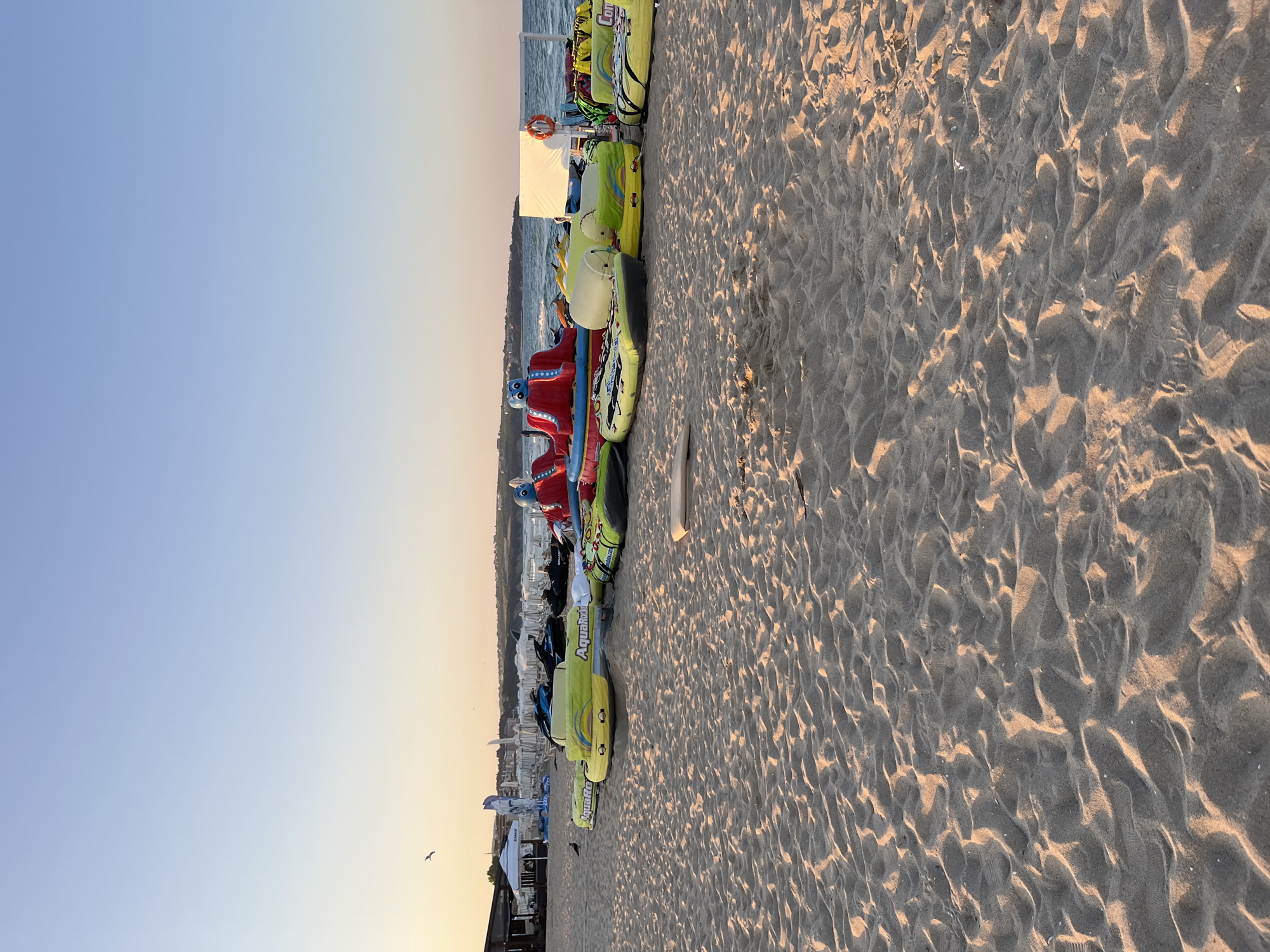 This is a wide-angle, eye-level shot of a sandy beach in Obzor, Bulgaria, during the early morning or late evening, as indicated by the warm, soft lighting and long shadows cast across the sand. The beach is lined with various inflatable water attractions, including what appear to be banana boats and towed tubes, in bright colors like yellow, green, and red. Some of these inflatables have branding visible, such as "AquaRocket." In the background, there are beach umbrellas, a building with a covered seating area, and a white structure with a life preserver. The sea is visible in the distance, calm under a clear, pale blue sky that transitions to a warmer hue near the horizon. A solitary bird is visible in flight in the upper left portion of the image, and another bird stands on the sand closer to the inflatables. The sand itself is textured with ripples and footprints, suggesting it is a public beach. The overall scene depicts a recreational area preparing for or winding down from a day's activities.
