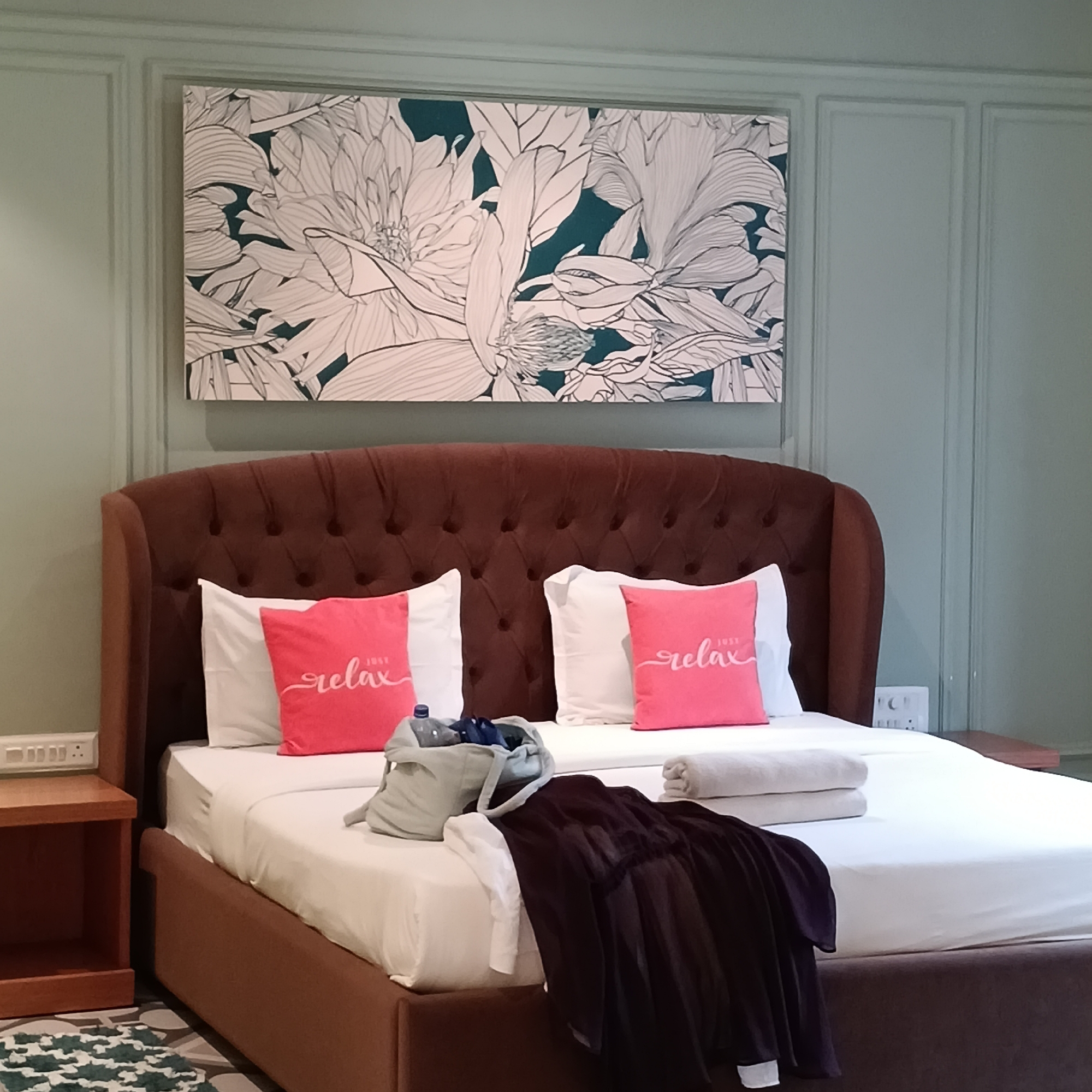 The image shows a hotel room interior. The room features a queen-sized bed with a brown upholstered headboard. The bed is made with white sheets and has two white pillows, each adorned with a small, bright pink decorative pillow that reads "Just relax".
A grey tote bag is placed on the bed, next to a stack of rolled-up towels. A dark-colored garment is draped over the lower portion of the bed.
Above the bed hangs a large rectangular piece of artwork featuring a floral design in white against a muted teal background. The wall is painted a light sage green, and has a paneled effect. A dark brown nightstand is visible on the left side of the bed, and a power outlet is noticeable on the wall behind the nightstand and beside the bed.
The location is identified as Calangute, India.