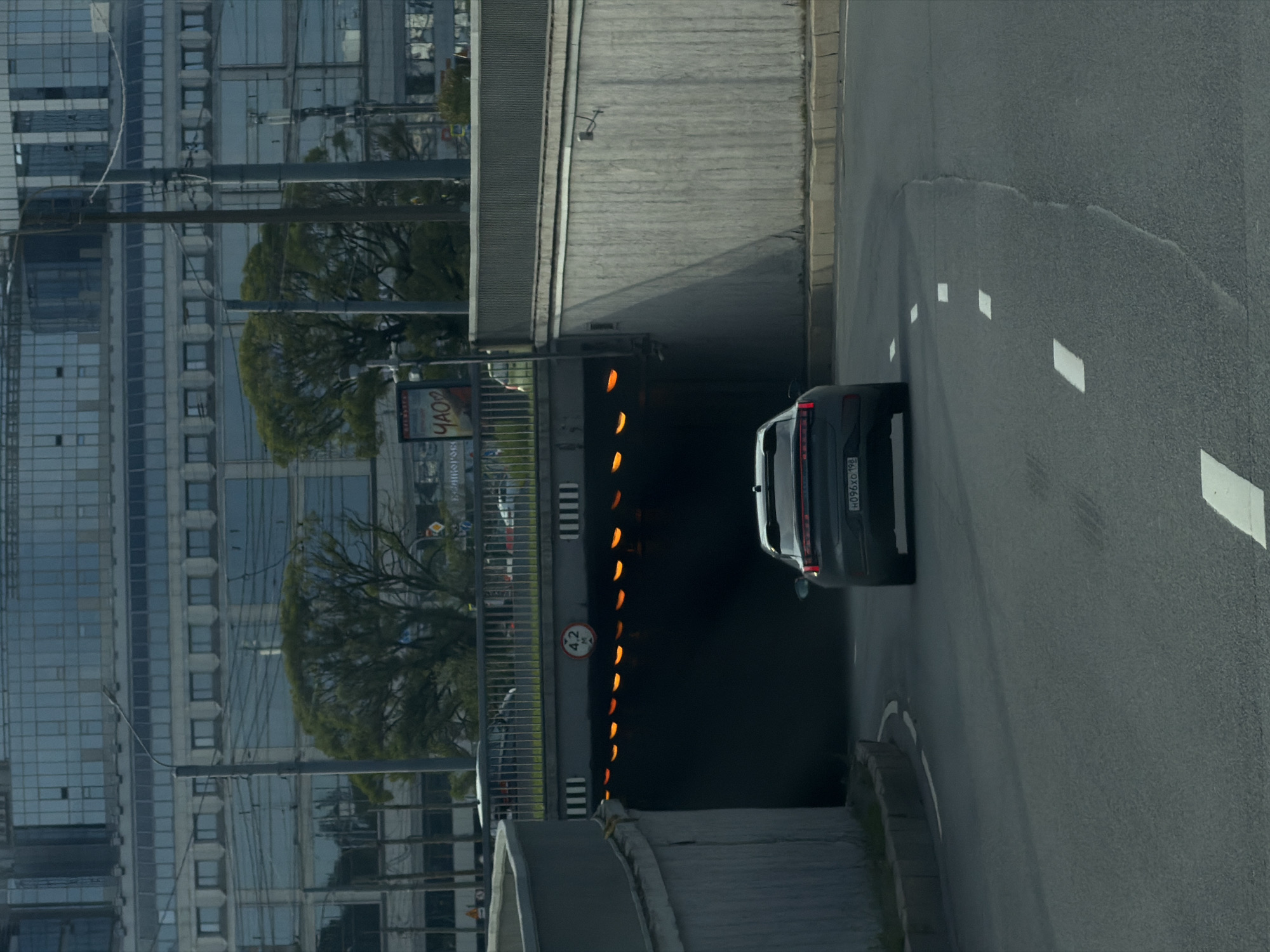 The image depicts a scene on an overcast day in Saint Petersburg, Russia, likely on an approach to an underpass. A dark-colored sedan with license plate "H096XO 198" is driving towards the viewer and entering a tunnel. The tunnel is illuminated by a series of orange lights, and a circular sign indicating a height limit of "4.2 M" is visible above the tunnel entrance.
The foreground consists of a two-lane asphalt road with white lane markings. The road curves slightly to the left as it approaches the underpass. The underpass itself is constructed of concrete, with textured walls. To the left, a grey concrete barrier wall lines the road.
In the background, behind the underpass, a large, modern multi-story building with a glass facade dominates the skyline. Several trees with green foliage are visible between the underpass and the building, and utility poles and wires crisscross the scene. A billboard with Russian text, including the word "ЧАСО" (Chaso), is also present on the left side of the image, near the overpass. The overall impression is an urban environment, possibly a transportation artery within the city.