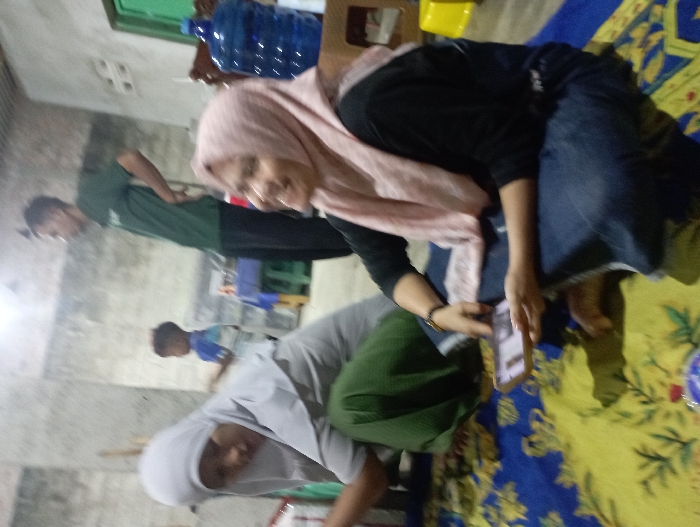 This image depicts an indoor scene, likely within a home in Pubian, Indonesia, featuring several individuals engaged in casual activities. In the foreground, a woman wearing a light pink hijab, a black long-sleeved top, and blue jeans is seated or crouched on a vibrant blue and yellow patterned mat. She is holding a smartphone in her hands, looking towards the viewer with a clear smile, suggesting a pleasant or joyful mood.

Behind her, to the left, another woman wearing a light gray or white hijab and dark green pants is seen bending forward, appearing to be focused on something on the floor or attending to an item. Further in the background, a man dressed in a dark green t-shirt and dark trousers stands upright, with his hands on his hips. A young boy in a blue and white shirt is partially visible behind the bending woman.

The setting includes plain, light-colored walls and a distinctive patterned floor mat, characteristic of a living or common area in a home. A large blue gallon-sized water bottle and a yellow plastic item are visible on a surface in the background, possibly a table or stool. The overall atmosphere is relaxed and informal, capturing a candid moment of daily life. The image is somewhat blurred, especially in the background. No discernible text is visible on any objects or screens within the frame.