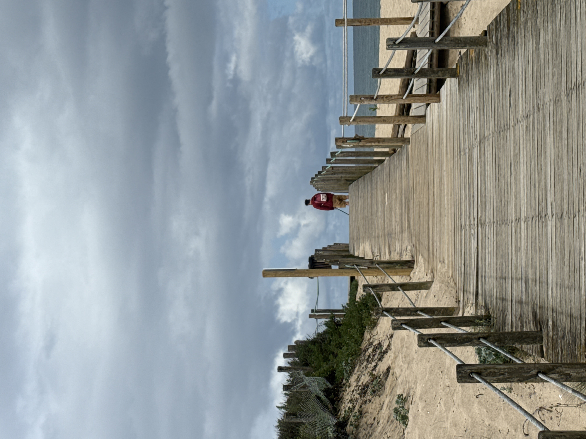 A wooden boardwalk stretches across the scene, leading towards a beach under a cloudy sky. A person in a red jacket walks on the boardwalk. The location is Vila Nova de Gaia, Portugal, evident from similar boardwalk structures along its coastline. The boardwalk is lined with wooden posts connected by cables acting as a railing, providing access to the beach. Sand and vegetation are visible on either side of the boardwalk. The sky is predominantly grey, suggesting overcast weather.