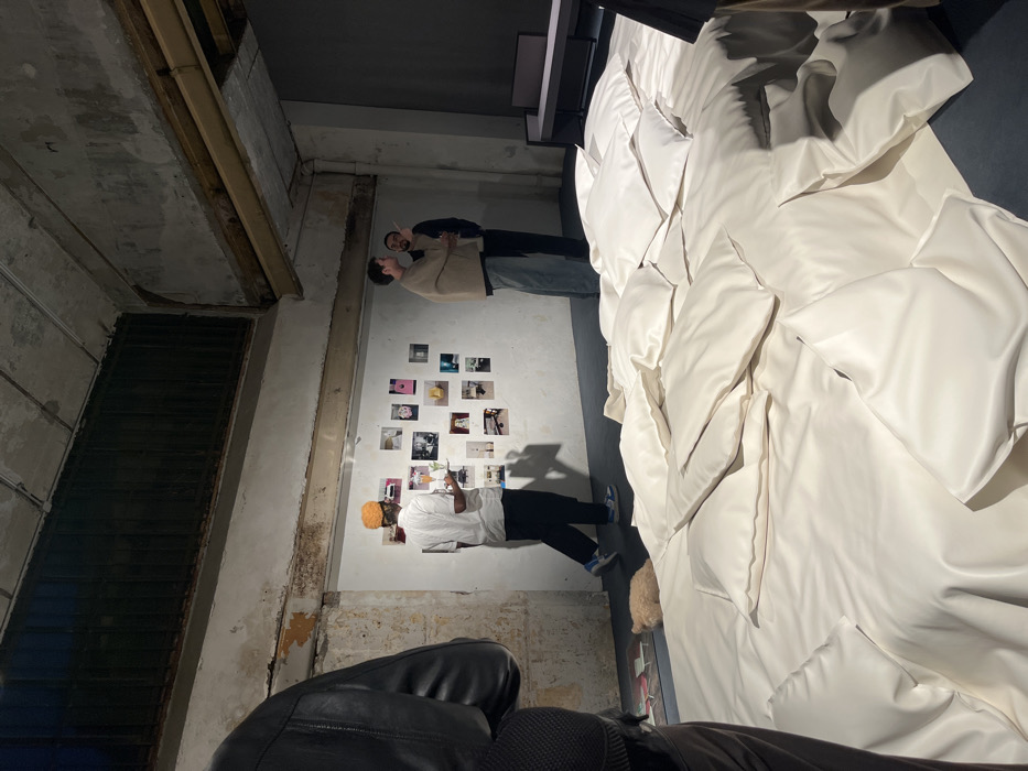 The scene depicts an interior view of an industrial-style building, likely repurposed for an exhibition or event, located in Paris, France. The space features rough, off-white concrete walls with distressed textures, a dark ceiling structure with visible beams, and a dark floor.
In the immediate foreground on the left, the back and left shoulder of an individual wearing a black leather jacket are visible. The central and right portions of the foreground are dominated by a large, floor-level art installation or display comprising numerous irregularly shaped, matte white soft forms resembling oversized cushions or pillows, arranged in a dense, contiguous mass.
In the mid-ground, a man wearing a white collared shirt, dark trousers, and an orange knit cap is seen walking from left to right, holding a small white object in his right hand. Behind him, an informal grid of approximately 15-20 small photographs or prints is affixed directly to a section of the white wall. Further right, two men are standing and interacting. One man, wearing a beige poncho-like garment, faces the second man, who is dressed in dark clothing and holds a thin, dark object near his mouth.
In the background on the far right, minimalist dark furniture, including a low bench or table with a light-colored top, is visible against a dark wall. A small, light brown, fuzzy object rests on the dark floor near the base of the white installation in the lower-left area.