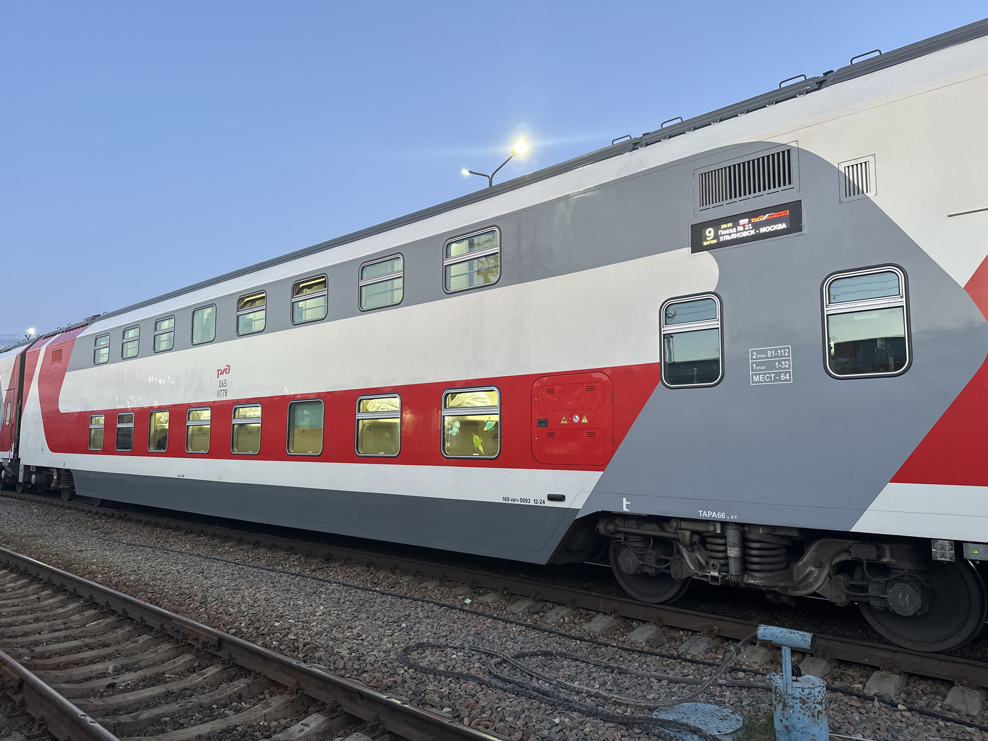 The image shows a modern, double-decker passenger train car parked on a railroad track. The car is painted in a color scheme of white, grey, and red. The destination sign indicates the train is number 9, traveling from Ulyanovsk to Moscow. Text on the side indicates the car is rated for 160 km/h. Some text appears to be in Russian. Through the windows, the silhouettes of passengers and their belongings are visible. The tracks are ballasted with gravel, and an industrial lamp post provides illumination. The sky is pale blue, suggesting early evening.