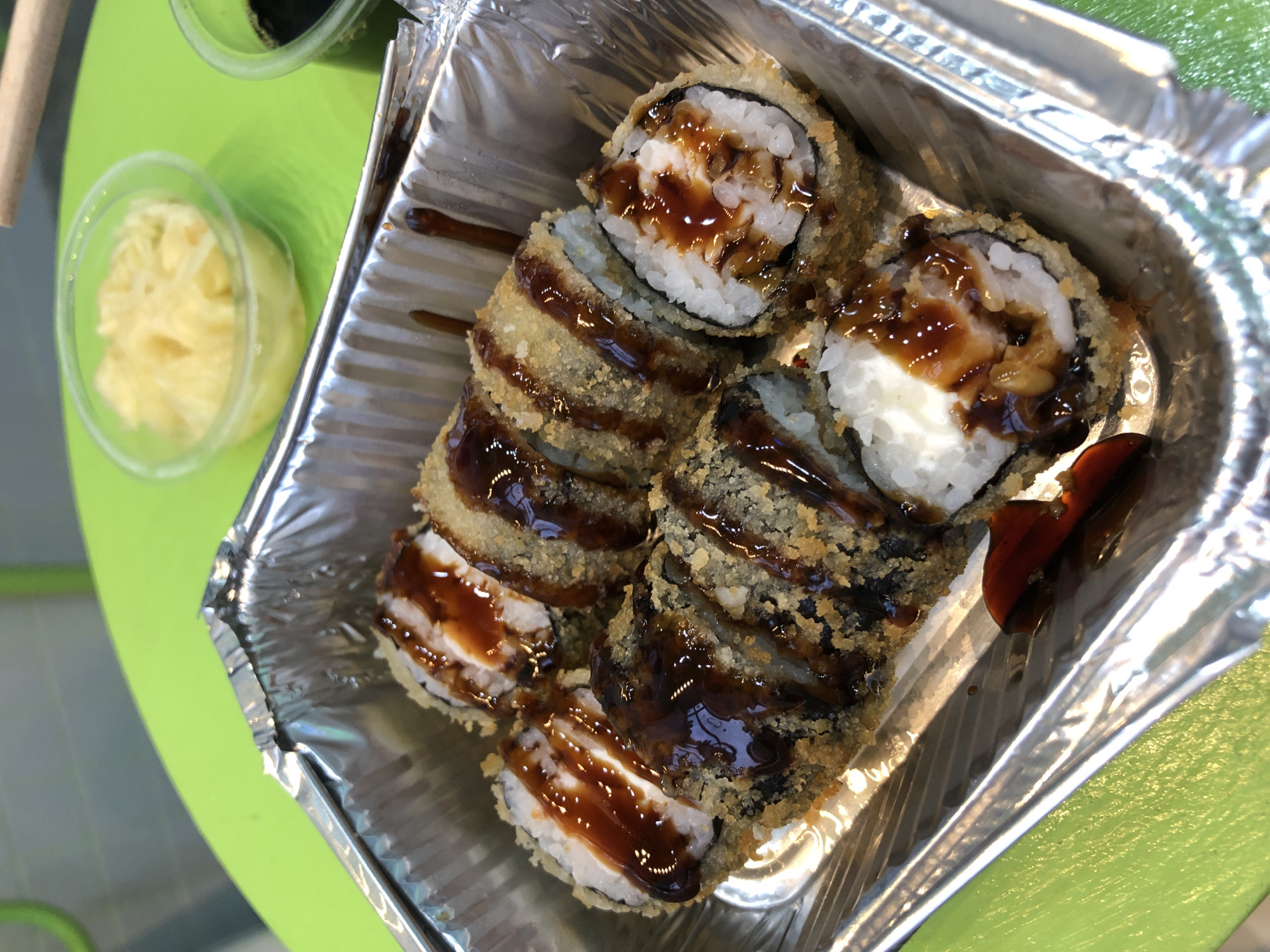 This media file shows a close-up, overhead view of a serving of sushi rolls in a foil container. The sushi rolls are fried and drizzled with a dark, glossy sauce, possibly teriyaki or eel sauce. The rolls appear to be filled with white rice and some form of white filling, possibly cream cheese or seafood. The exterior of the rolls is coated in breadcrumbs or tempura batter, giving them a crispy, golden-brown appearance.
In the background, to the left of the container, are two small plastic containers. One contains a pale yellow, shredded ingredient, likely pickled ginger. The other, partially visible, appears to contain a dark liquid, possibly soy sauce. The setting appears to be a casual dining environment, with a bright green surface beneath the food container, suggesting a table or counter. The lighting suggests it could be daytime or under bright artificial lights. No people are visible in the frame. The overall presentation suggests a take-out or casual dining experience. There is no visible text in the image.