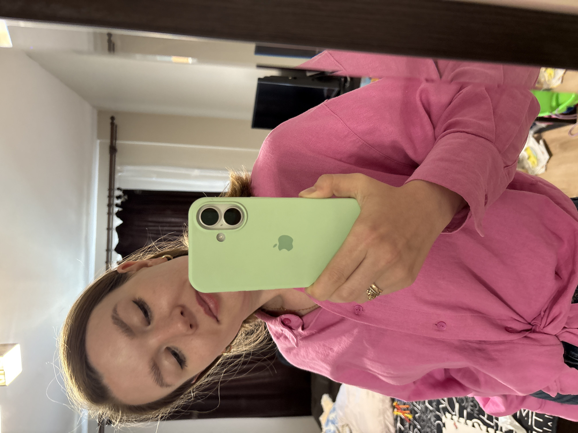 A woman with light brown hair is taking a selfie in a mirror. She is wearing a pink button-down shirt. She is holding a light green iPhone in front of her face. The background shows a room interior with a television and other furniture, and drapes. The location is potentially Izhevsk, Russia, given the request.