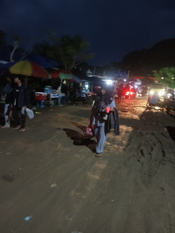The image depicts an outdoor night scene in Kecamatan Pujut, Indonesia, resembling a lively market or gathering area. The sky is dark, characteristic of nighttime, with various artificial lights illuminating the sandy, uneven ground.
In the foreground, a woman, identifiable by her hijab and light-colored attire (possibly a light blue tracksuit), walks from left to right. She holds a red cup, a dark jacket is draped over her arm, and she carries a pink bag. A distinct shadow stretches behind her. To her left, slightly further back, a man in a dark jacket and light pants walks away from the viewer.
The background is filled with informal market stalls, distinguished by colorful umbrellas in shades of red, yellow, blue, and green. These stalls are brightly lit, showing details like coolers and plastic tables. Several people are gathered around these stalls. On the right side of the frame, multiple motorbikes or scooters are present, their headlights and taillights creating bright streaks of light. One vehicle appears to be a three-wheeled transport. Faint outlines of hills are visible against the dark horizon. No visible text is discernible.
