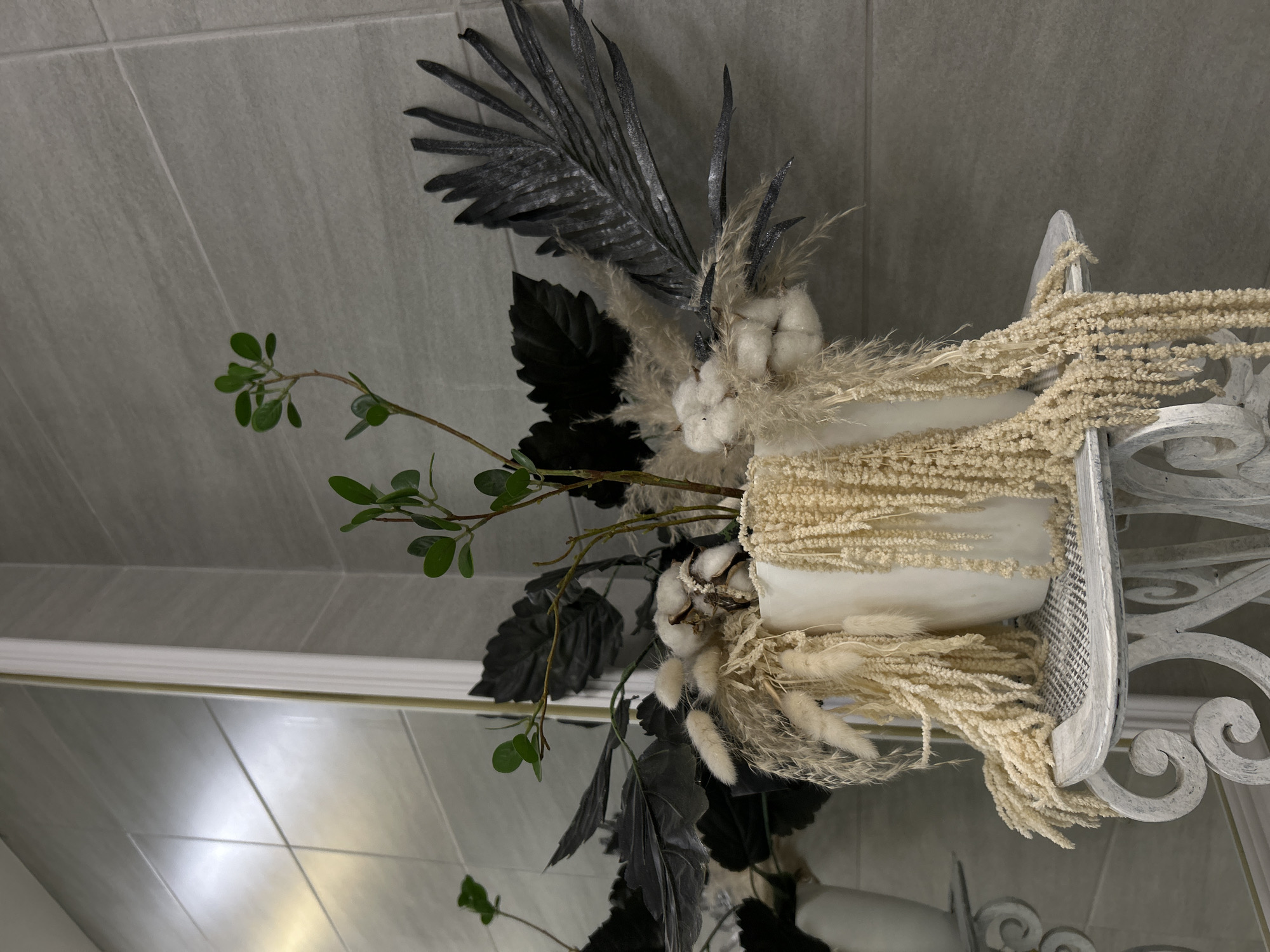 A decorative arrangement is set against a neutral grey tile wall and reflected in a mirror below. The arrangement includes a white vase within a distressed white metal stand featuring scrollwork. The vase holds a variety of floral elements, including dried cream-colored grains, cotton bolls, black painted leaves, black painted feather-like branches, and green leafy branches. The reflection in the mirror shows the lower part of the arrangement and the tile wall. The lack of typical markers prevents determining whether this is Saint Petersburg, Russia, or any other location.