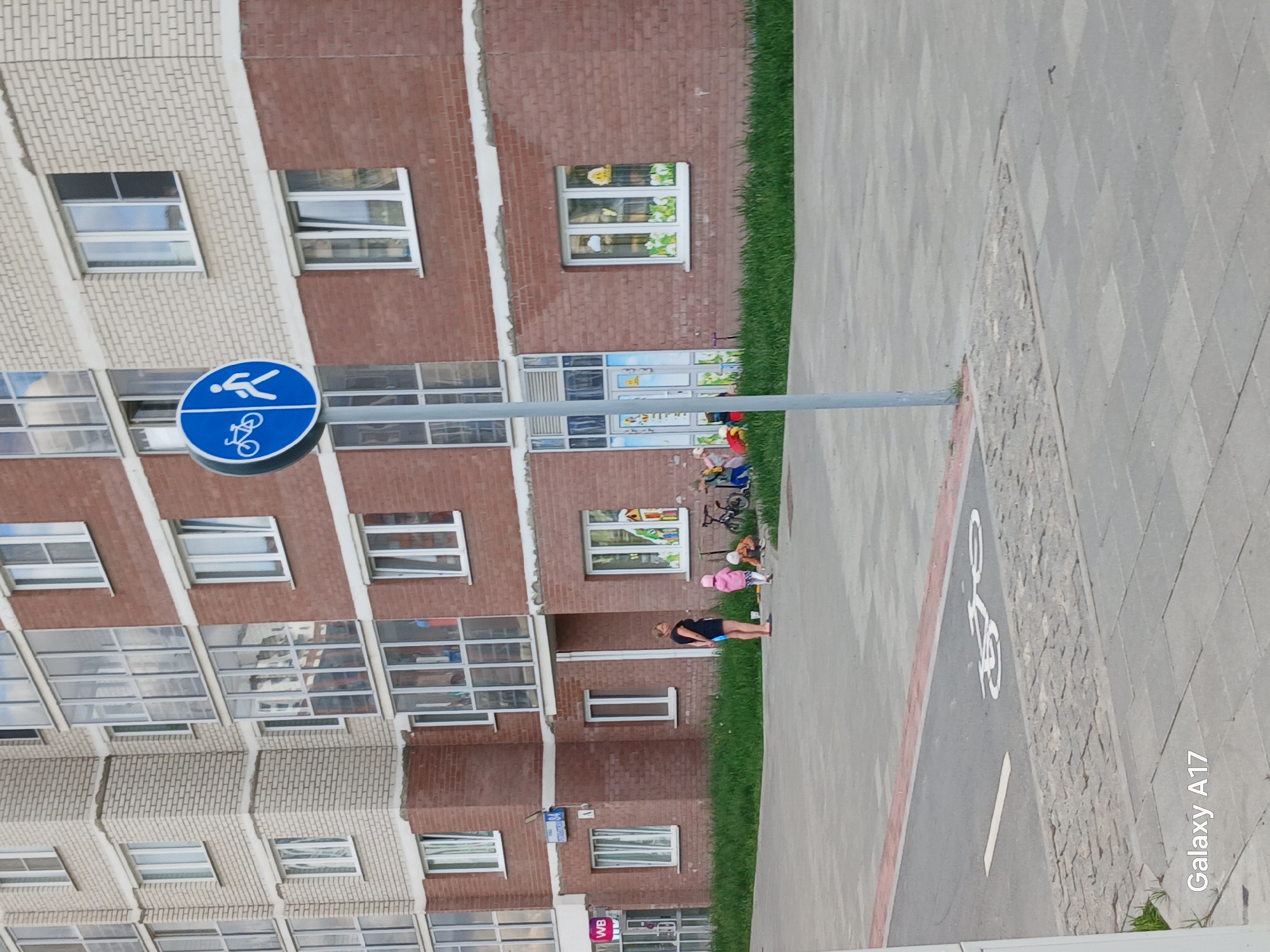 The image captures an outdoor scene in a residential area during what appears to be a clear, sunny day. The background is dominated by an apartment building constructed with alternating sections of light-colored and dark red brickwork. Numerous windows are visible across the facade, some reflecting the sky and surroundings, while others on a lower floor of the red brick section are adorned with child-friendly decorations like a yellow sun and green floral motifs, suggesting a children's facility or residential unit.
In the foreground, a paved pathway runs parallel to the building. This path features distinct sections: a lighter-colored pedestrian area and an adjacent darker grey strip designated as a cycle path. The cycle path is marked with a white bicycle symbol painted on its surface and delineated by a red stripe. A blue circular sign, mounted on a metal pole, stands prominently; it displays white pictograms for both a pedestrian and a bicycle, indicating a shared path.
On a strip of green grass situated between the building and the paved area, an adult woman, dressed in a dark top and shorts, stands. Nearby, two small children are visible, one wearing a pink hat, appearing to be playing or resting on the ground amidst some toys or belongings. Another adult or older child and a bicycle are partially seen further along the grass strip.
Visible text includes a "Galaxy A17" watermark in the bottom right corner of the original image orientation. On the building facade, a white sign with a "WB" logo, likely for Wildberries, is visible. Additionally, a blue sign with the number "1" is partially discernible on the building. The scene portrays a typical peaceful outdoor environment, with residents and children utilizing shared spaces.