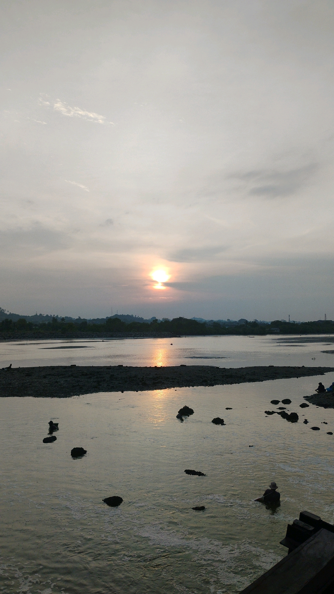 The image depicts a serene sunset over a body of water, likely a river or a bay, in Lhokseumawe, Indonesia. The sun, a bright orb, is partially obscured by hazy clouds, casting a warm, golden reflection on the water's surface. The shoreline is characterized by a rocky or sandy bank on the left, transitioning into a darker landmass with scattered trees and structures in the distance.
In the foreground, several dark, irregularly shaped rocks emerge from the shallow water. One or two individuals appear to be partially submerged in the water, perhaps fishing or bathing, with one person on the right side of the frame visible from the waist up, wearing a hat. On the far right, a dark, wooden structure, possibly a pier or a jetty, is partially visible. The overall atmosphere is tranquil, with the muted colors of the sunset and the calm water suggesting a peaceful evening. The presence of natural elements like water and rocks, combined with the distant hint of civilization, creates a harmonious landscape.