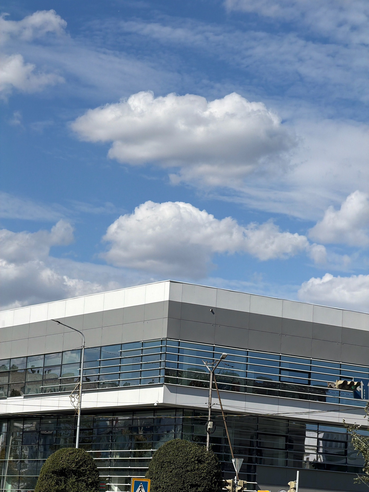 The image shows the exterior of a modern building with large glass windows, reflecting the sky and clouds. The building has a gray and white facade. In the foreground, there are manicured bushes and a street lamp. Traffic lights and pedestrian crossing signs are visible, suggesting an urban environment. The sky is blue with white, fluffy clouds. This image was captured in the city of Pavlodar, Kazakhstan.