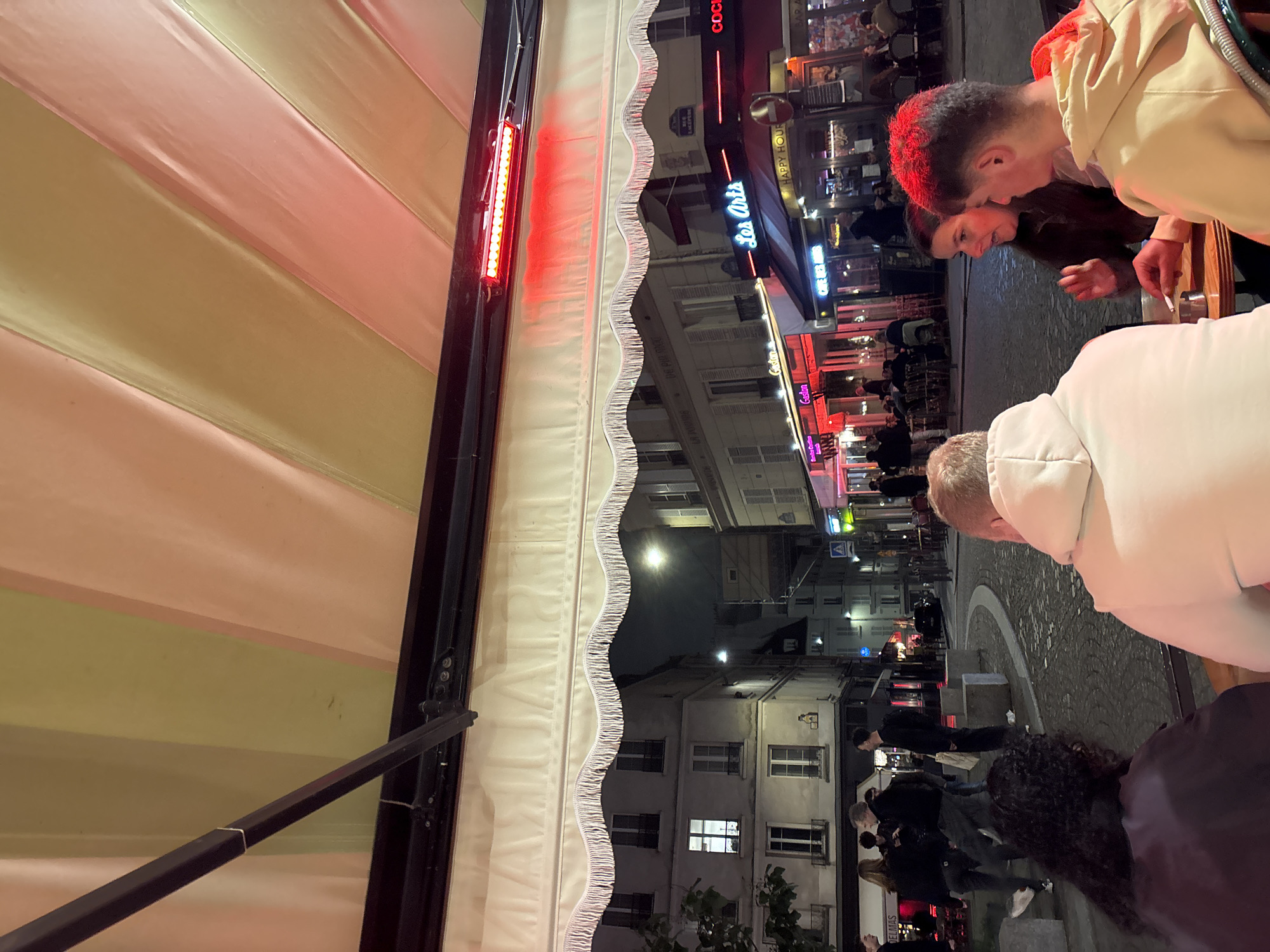 The image shows a night scene from an outdoor seating area, likely a café or restaurant in Paris, France. The foreground features part of a striped awning with a decorative fringe and a red light fixture attached to its side. Several people are partially visible; one wears a white hooded sweatshirt, and a couple stands together with the male wearing a light-colored hooded sweatshirt. They appear to be on a cobblestone street lined with buildings, many of which have glowing signs in French such as "Les Arts" and "Gadjon." The street is busy with pedestrians, and lights illuminate the buildings and streets, creating a vibrant urban ambiance.