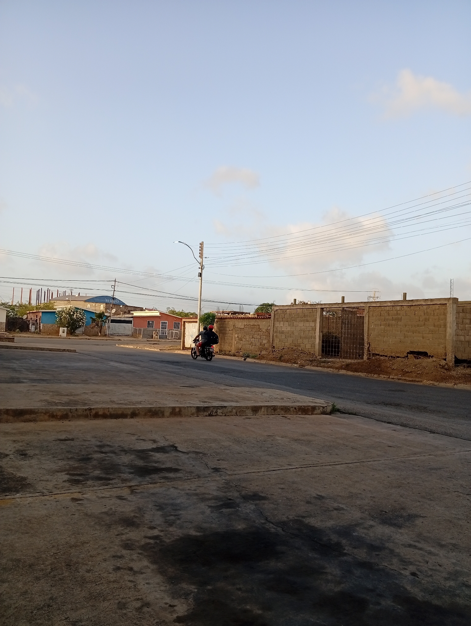 This image depicts an outdoor street scene in Juan Griego, Venezuela, under a bright, light blue sky with scattered wispy clouds, suggesting either early morning or late afternoon. The foreground features a concrete or paved surface with dark, wet-looking patches.
A person wearing dark clothing and a red helmet is visible riding a motorcycle away from the viewer on a paved road that curves through the middle ground.
On the left side of the road, several buildings of varying styles and colors are present. One notable structure is painted light blue and has partially visible white text that appears to read "Colegio" (school) or similar. Another building is pink, and further in the background, a structure appears to be under construction with exposed rebar. Sparse green foliage is scattered among these buildings.
To the right of the road, a long, low wall constructed from concrete blocks runs parallel to the street, featuring a metal gate or opening. Utility poles with multiple overhead power lines traverse the scene against the sky. The overall scene conveys a quiet, everyday street environment in a residential or mixed-use area.
