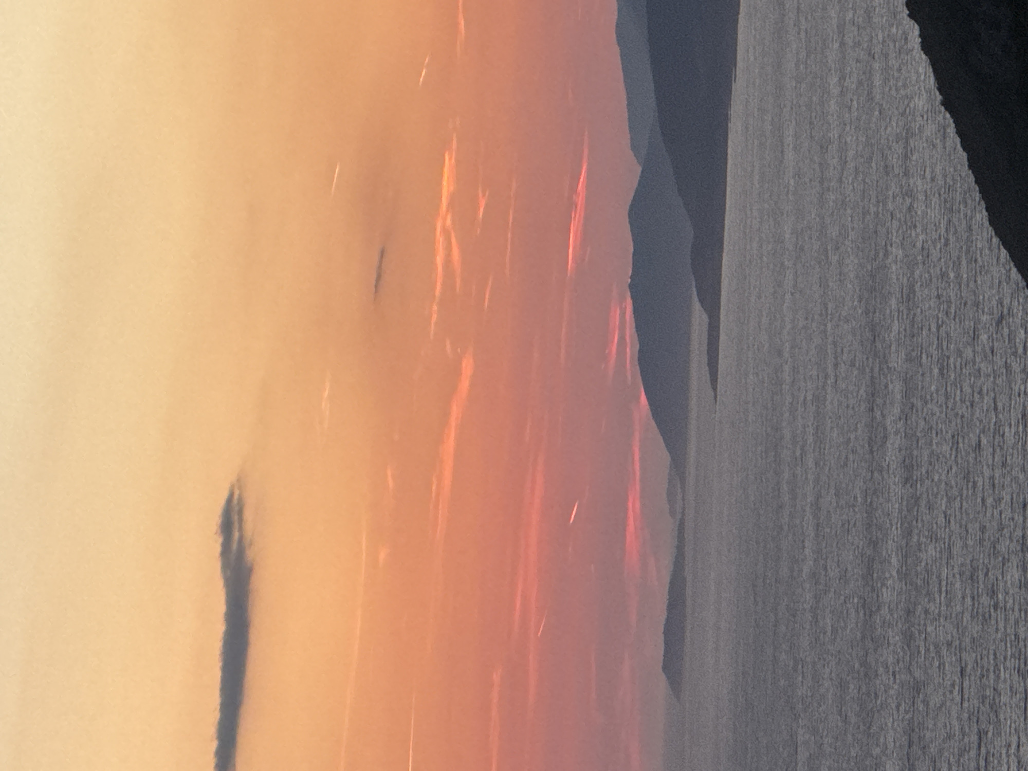 This is an eye-level, medium shot photograph of a sunset over the Adriatic Sea, likely near Zagora, Montenegro. The sky is a gradient of warm colors, transitioning from pale yellow at the top to vibrant orange and pink hues lower down. A single, elongated dark cloud is visible in the upper left quadrant. Streaks of contrails from aircraft are faintly discernible in the sky. The sea occupies the lower half of the frame, appearing calm with a textured surface. In the distance, a series of silhouetted, undulating islands or coastal landmasses are visible against the horizon. The foreground features a dark, sloped landmass on the bottom right, suggesting a viewpoint from a hill or cliff overlooking the sea. There are no people or visible objects in the scene other than natural elements. The overall impression is one of serene natural beauty during twilight.