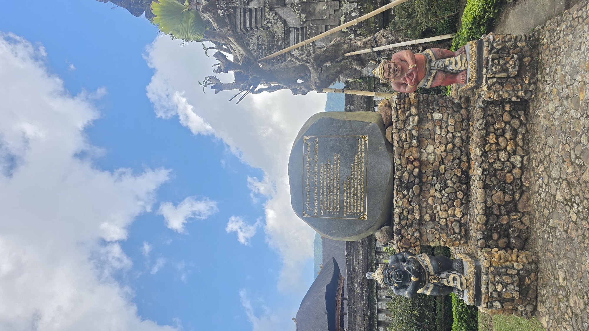 This image, shot outdoors in Baturiti, Indonesia, showcases a stone sign etched with rules for visiting the "PURA PENATARAN AGUNG ULUN DANU BERATAN". The sky, a blend of blue and white clouds, serves as the backdrop for the sign, which is positioned between two stone statues. The statue to the right, with reddish skin, is a reclining figure while the one on the bottom is sitting. The statues are mounted on stone steps. A portion of a building with a peaked roof is seen in the background, along with a tree with bare branches. The stone sign contains text in both Indonesian and English, providing guidelines for tourists visiting the temple area.