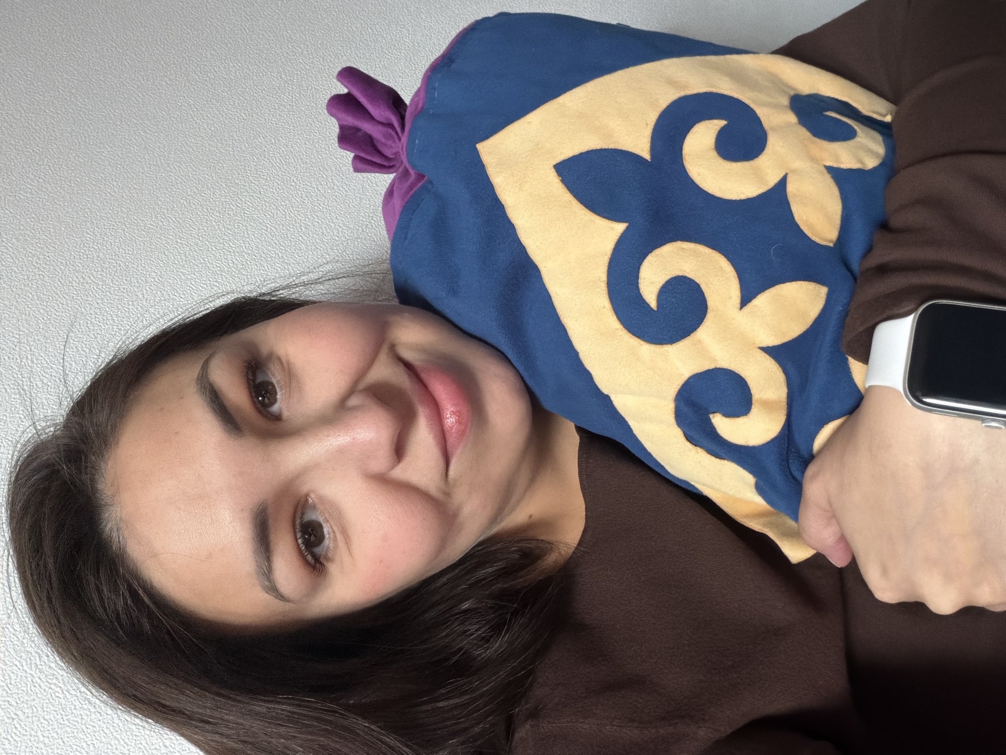 An indoor, close-up, rotated selfie shows a young woman with a gentle smile, positioned in the lower-left portion of the frame. She has long dark hair, brown eyes, and is looking directly at the camera. Her complexion is fair, and she appears to be in her 20s or 30s.
The woman is wearing a dark brown, long-sleeved top. On her left wrist, a white smartwatch with a dark screen is visible. She is holding or resting her head against a distinctively patterned pillow or cushion. The pillow is predominantly blue with a large, intricate, gold or tan-colored traditional design or emblem. The top of the pillow is cinched and made of a purple fabric.
The background is a plain, light-colored (likely white or light grey) wall with a subtle, textured surface. The overall impression is one of calm or contentment. No discernible text is visible on any object or clothing. This scene takes place in Korolyov, Russia.
