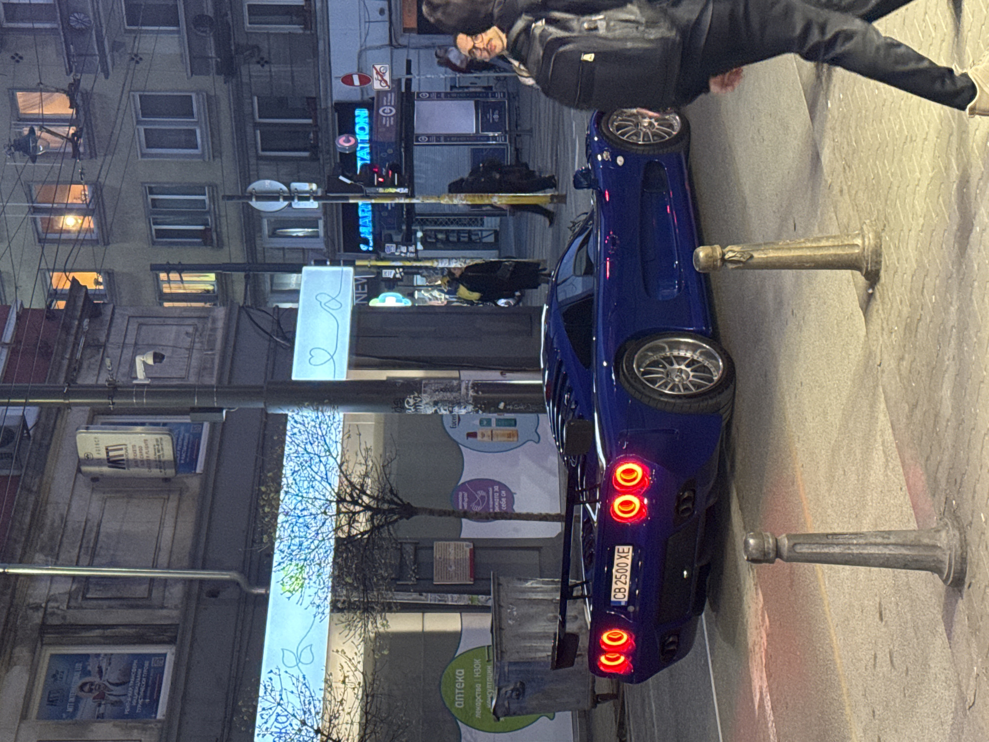 A vertical street-level shot shows a blue sports car parked on a sidewalk in Sofia, Bulgaria. The car's license plate, "CB 2500 XE", is visible. A person with a backpack is bent over near the car, their face obscured. In the background, there are buildings with lit windows and storefronts, some with visible signs. One sign reads "TATTOO", another "MAR STATION", and there's a pharmacy sign in Bulgarian. Streetlights and utility lines are also visible. The sidewalk has metal bollards along the edge.