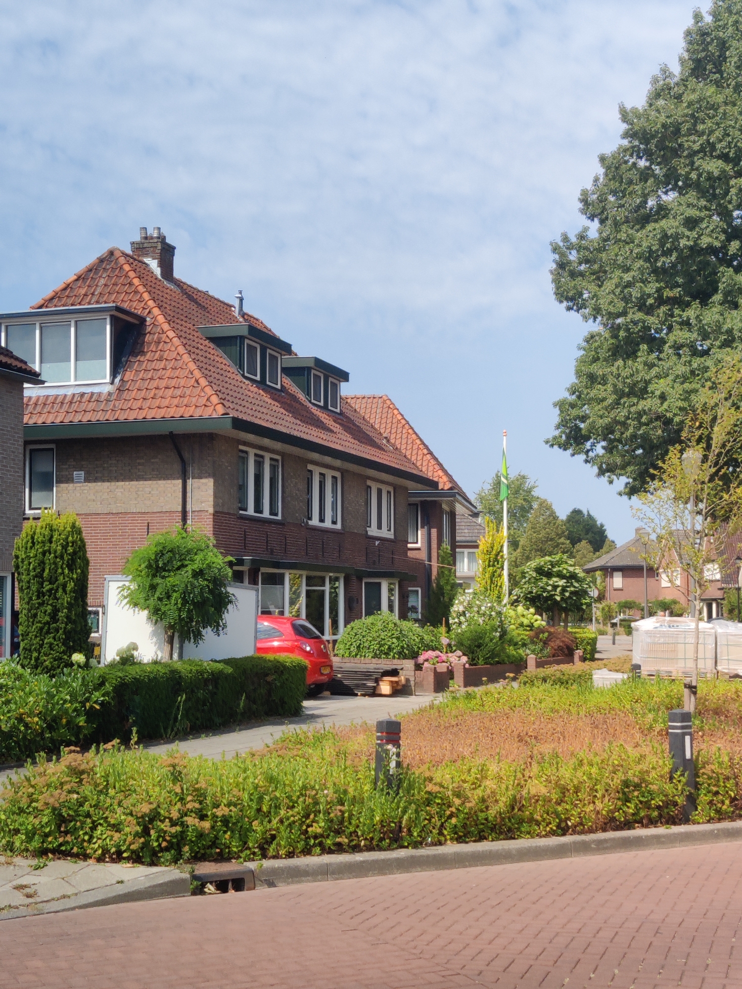 The image depicts a suburban street scene in Winterswijk, Netherlands. In the foreground, a red car is parked in front of a brick house with a red tile roof. The house has several windows and dormers. Adjacent to the house is a well-maintained garden with green bushes and flowering plants. In the background, more houses and trees are visible under a bright, partly cloudy sky, suggesting it is daytime. A flag, green and white, flies from a flagpole in the distance. The overall impression is one of a peaceful, residential neighborhood on a pleasant day. There is no visible text in the image.