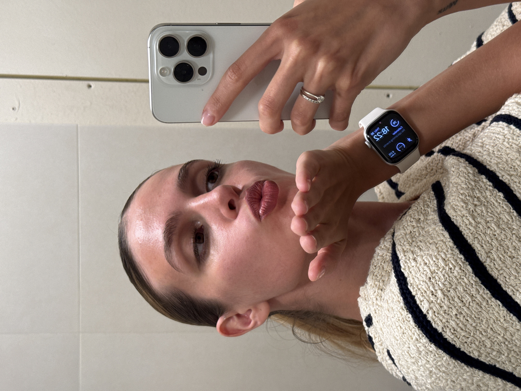 A woman is captured in a mirror selfie, seen from the chest up, with her head tilted. She has light skin, dark eyebrows, and her lips are pursed in a "kissing" pose, covered in a dark rose or berry-toned lipstick. Her light brown hair is visible around her ears and shoulders. She wears a textured, off-white or beige sweater with thick, dark horizontal stripes.
Her right hand holds a light-colored smartphone with a prominent square camera module housing multiple lenses (at least three large dark ones and one smaller lens/sensor), reflecting in the mirror. On her right ring finger, she wears two rings, one with small stones and another plain band. Her left arm is raised, and on her wrist is a white-banded smartwatch. The smartwatch screen, reflected in the mirror, clearly displays the time as "18:55" and various app icons, including activity rings, a moon icon (likely for focus/sleep mode), and a steps counter. Reversed text "dialsneIC" and "STneve gnimoC" can be seen, which would be "Calendar" and "Coming events" respectively. Faintly visible on her upper left arm is a reversed tattoo that appears to read "aetix".
The background is a plain, light-colored wall, possibly tiled, with a horizontal seam or line and some minor surface imperfections. The overall scene suggests an indoor setting. The time displayed on the watch indicates it is late afternoon or early evening. The woman's pose conveys a playful or confident mood. The photo is taken in Tbilisi, Georgia.