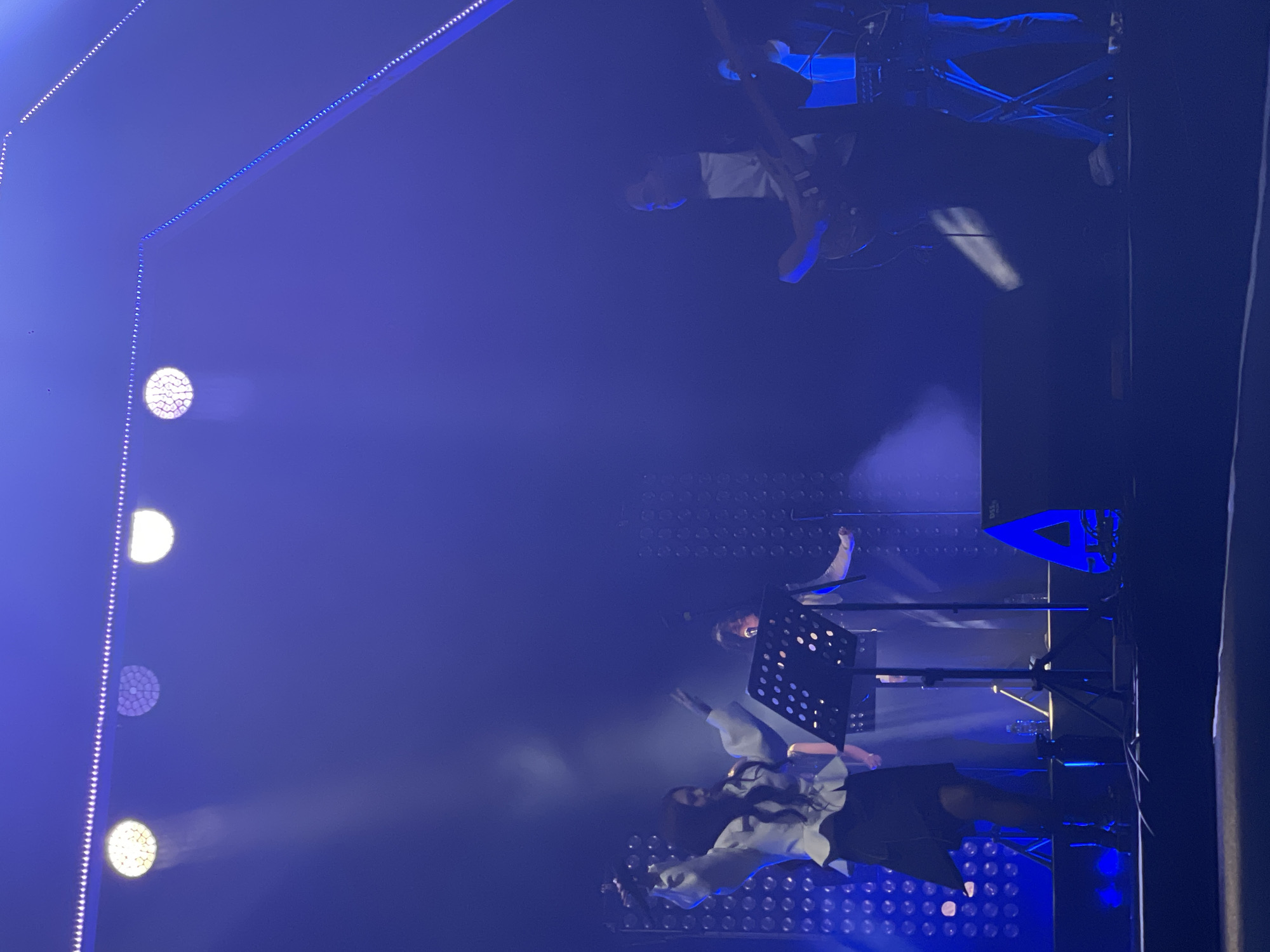 A rotated image of a dimly lit stage in what appears to be an indoor venue. Blue lighting dominates the scene. From left to right: a vertical series of circular stage lights, a person (presumably a singer) behind a music stand, a person on a higher platform with a music stand, and a guitarist on the upper right. The background appears to be an LED light wall. No specific details point to the city of Salembaran, Indonesia.