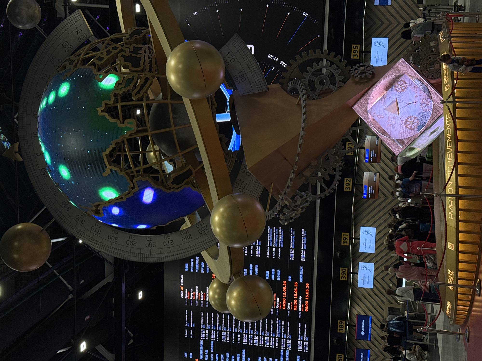 The image depicts an interior scene, likely within an airport terminal in Khimki, Russia. The focus is on a complex, ornate art installation comprising a globe with illuminated landmasses, several golden spheres, gears, and architectural elements like a compass on an overhead cube.
Below the artwork, there are multiple departure screens displaying flight information in both Russian and Chinese, with details like destinations (e.g., Yerevan, Chelyabinsk, Sochi), flight numbers, and registration times.
To the right of the screens, a queue of people is waiting behind a red rope barrier marked "AVIATION ACHIEVEMENTS" in Cyrillic and English. The waiting area has numbered markers from 388 to 395. The environment suggests a public space within an airport, possibly dedicated to showcasing aviation history or technological advancements.