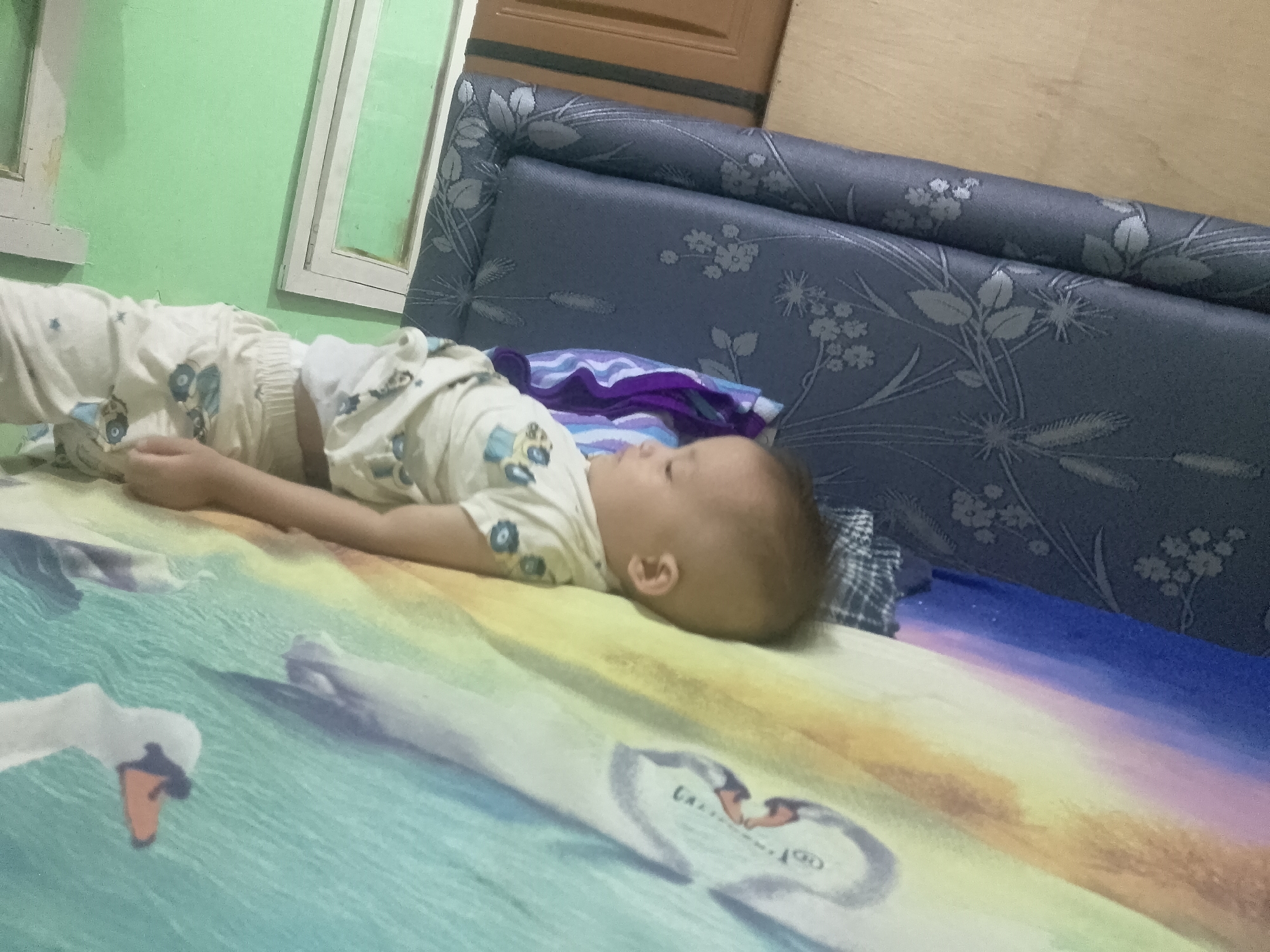 An infant is lying on a bed covered with a colorful sheet featuring swan designs. The infant is wearing a white outfit with a blue floral pattern. To the left of the infant is a green wall with a white window frame. Behind the infant is a blue headboard with a floral pattern. The scene appears to be in a bedroom in Cikalongkulon, Indonesia.