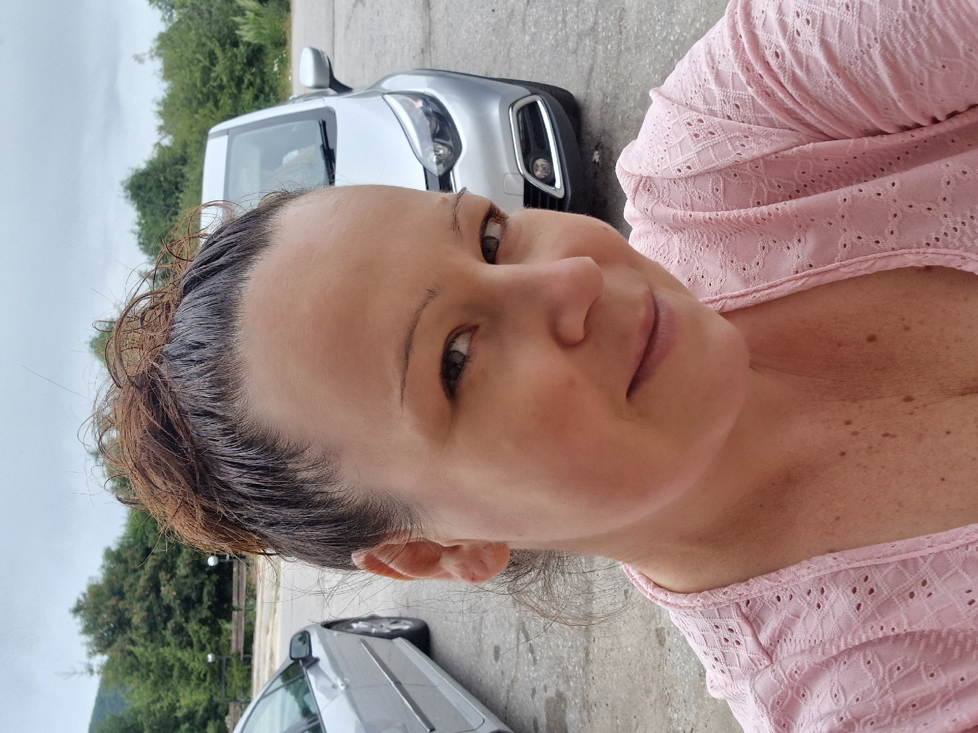 A woman is posing for a selfie outdoors. She has her hair pulled up in a messy bun, and she is wearing a pink top. In the background, there are two cars and some trees. The scene appears to be a parking lot in a natural setting, possibly near a mountainous area, given the presence of trees and a cloudy sky. The location is identified as Shipka, Bulgaria.