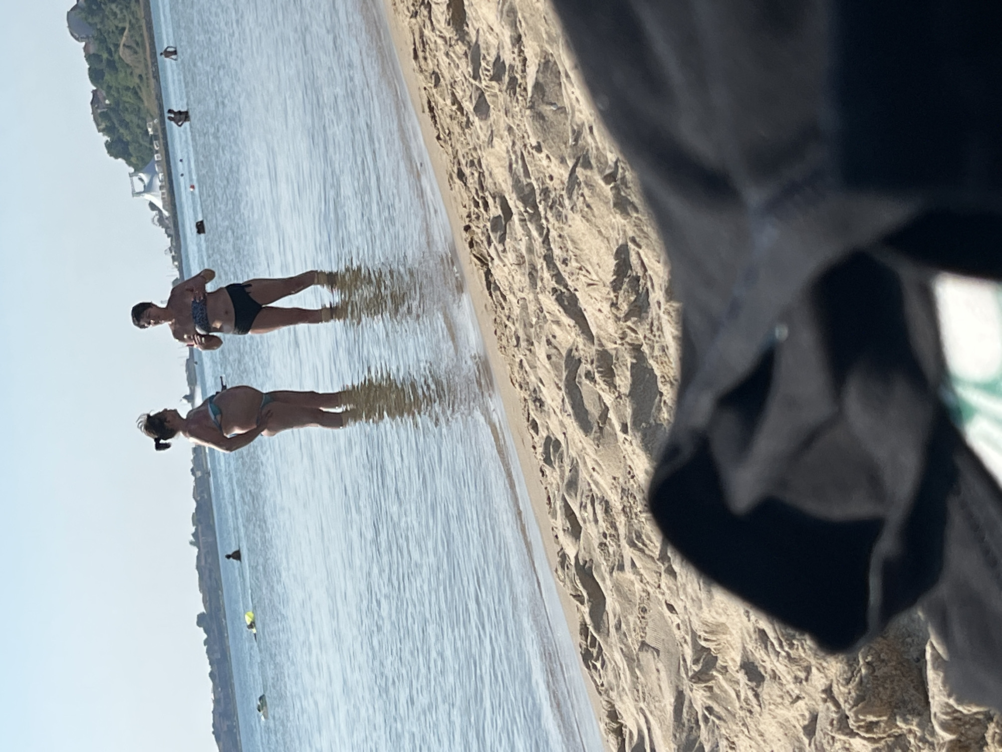 Two women are standing in shallow water on a sandy beach. The woman on the left is wearing a light blue bikini and appears to be pregnant. The woman on the right is wearing a patterned bikini top and black bikini bottoms. They are facing each other and appear to be conversing. In the background, there are several other people in the water and a few small yellow inflatables. Further in the distance, there is a coastline with buildings and a large white tent-like structure on a hill. The sky is clear and blue, and the sun is shining. The image appears to have been taken at a beach in Nesebar, Bulgaria, likely on a sunny day.