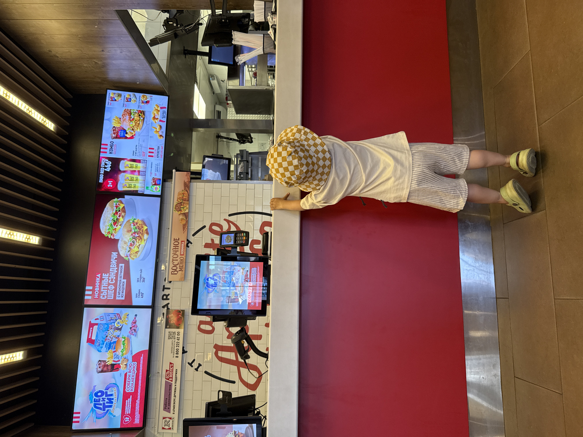 In Заволжье, Russia, a young child wearing a checkered bucket hat and a white t-shirt with striped shorts stands at a red counter in what appears to be a fast-food restaurant. The child is facing away from the camera, looking towards the counter. On the counter, there are two computer monitors displaying menu items and a credit card machine. Behind the counter, a kitchen area is visible with more equipment and monitors. The restaurant interior features a modern design with a wooden ceiling and lighting. The menu boards on the wall display various food and drink options, including sandwiches and combo meals, with Russian text. The overall scene depicts a typical fast-food restaurant setting with a child waiting at the counter.