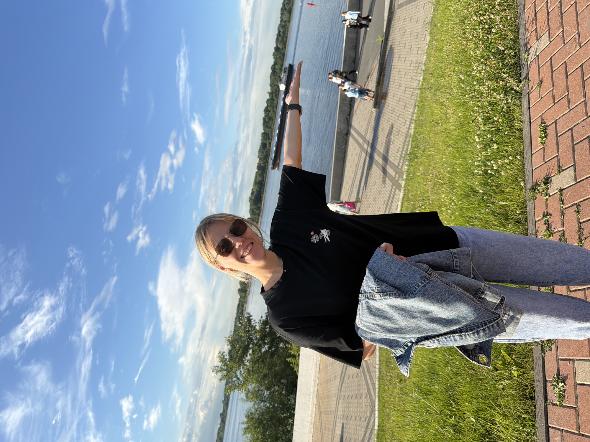 A young woman with blonde hair and sunglasses smiles as she poses for a photo. She is wearing a black t-shirt and blue jeans, with a denim jacket draped over her lap. Her right arm is raised, and her hand is pointed upwards. The background shows a wide river with a barge on it and trees lining the far bank. To the right, there is a paved walkway next to a grassy area with clover flowers, leading to a brick pathway. The scene appears to be outdoors on a sunny day with a bright blue sky filled with scattered white clouds. People can be seen walking along the pathway in the distance. The image is taken from a low angle, looking up towards the woman and the sky. The location context provided is Nizhny Novgorod, Russia.