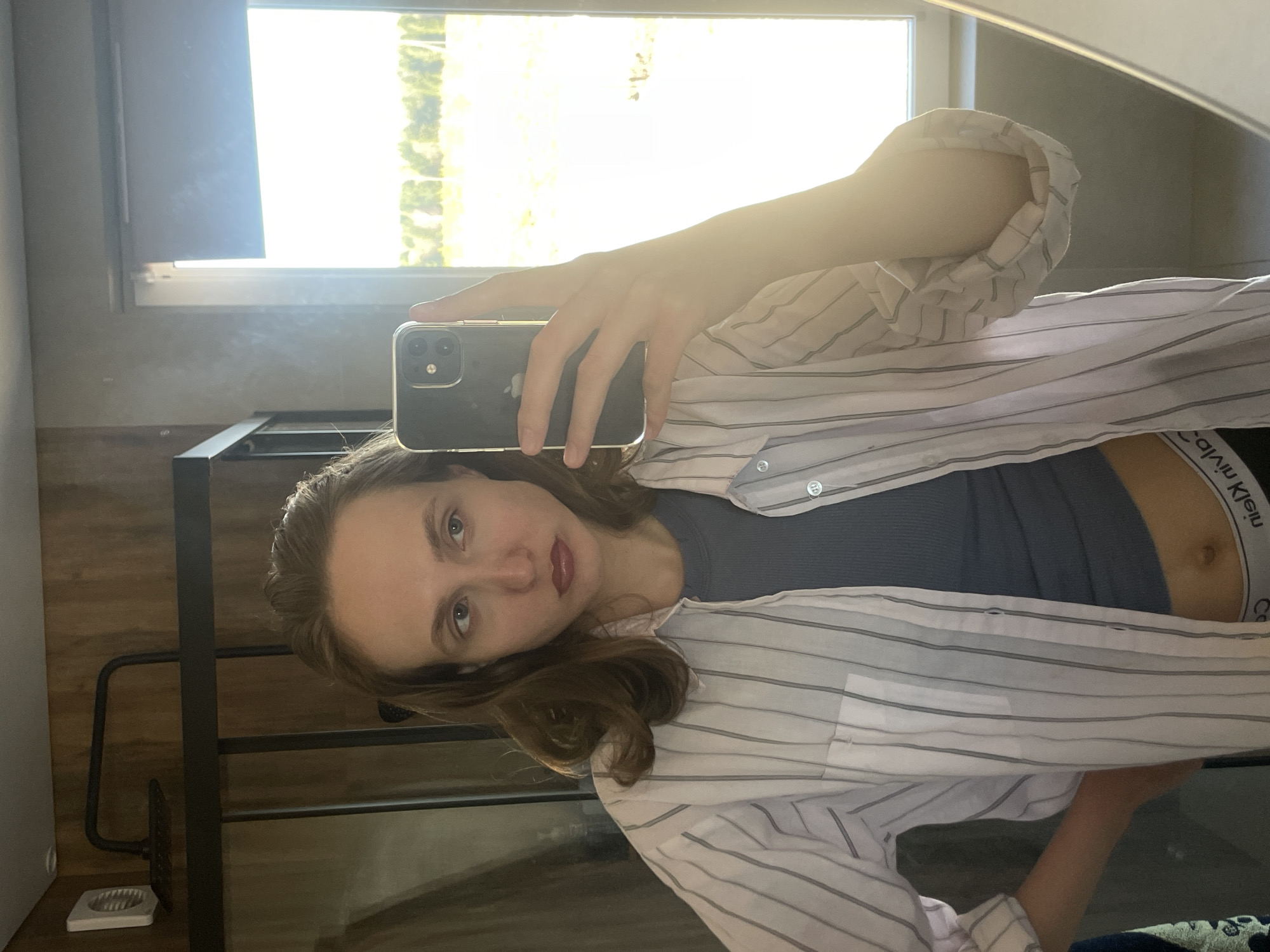 A woman takes a selfie in front of a mirror. She is wearing a dark gray top, a striped white and gray button-down shirt, and Calvin Klein underwear. She is holding a smartphone in her hand, partially obscuring her face. The background features a window with bright sunlight and a metal shelf with a white object on the floor next to it. The location, Подшивалово, Russia, cannot be verified from the image.