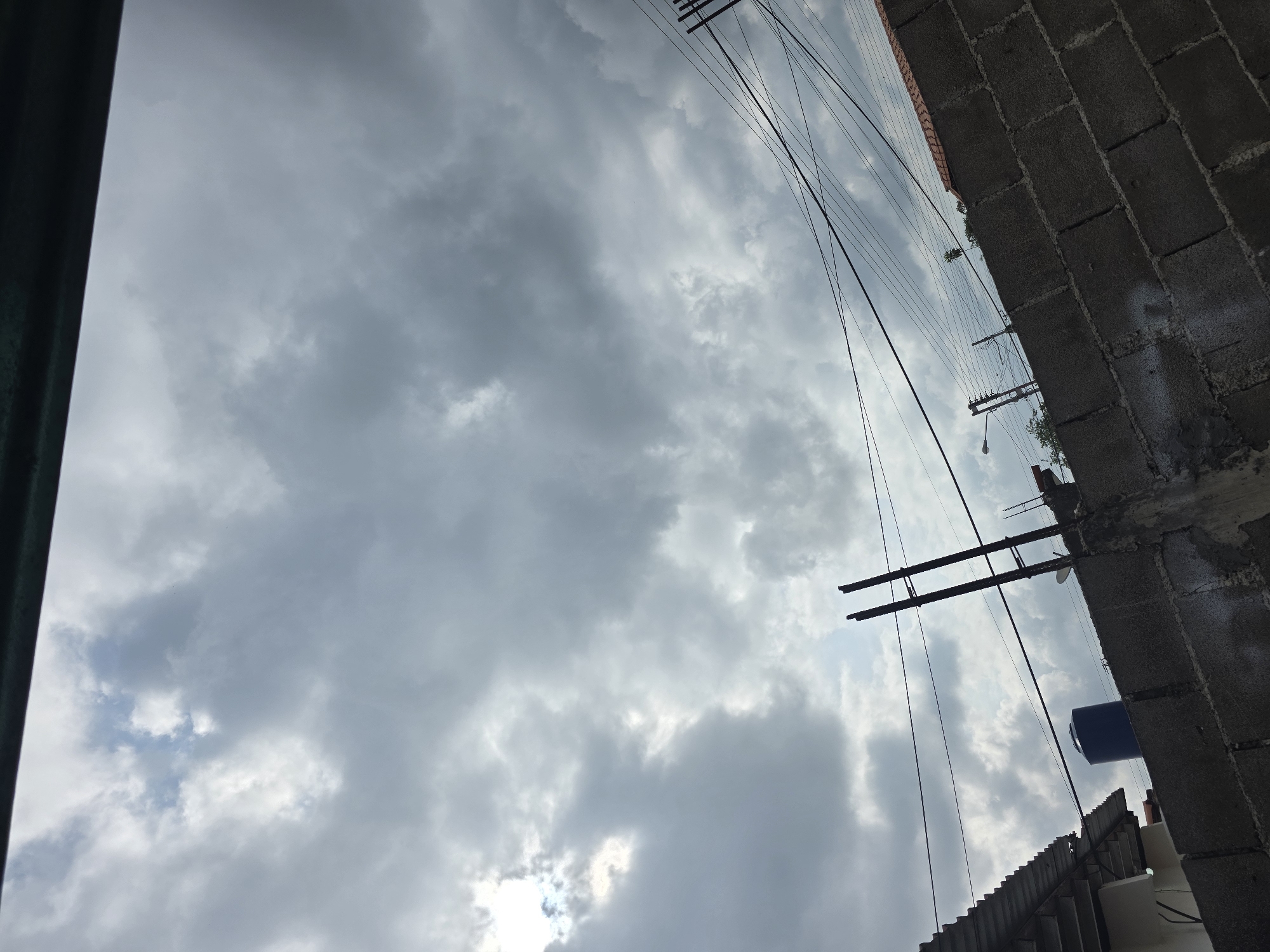 A low-angle shot captures a cloudy sky above Parroquia Ocumare, Venezuela. The foreground is defined by a rough, gray concrete block wall on the right and a dark green vertical structure on the left. Electrical wires and poles extend diagonally across the frame against the backdrop of the clouded sky. Below the wall, parts of a corrugated roof and a blue cylindrical tank are visible. The overall scene conveys a sense of urban infrastructure juxtaposed with a cloudy atmosphere.