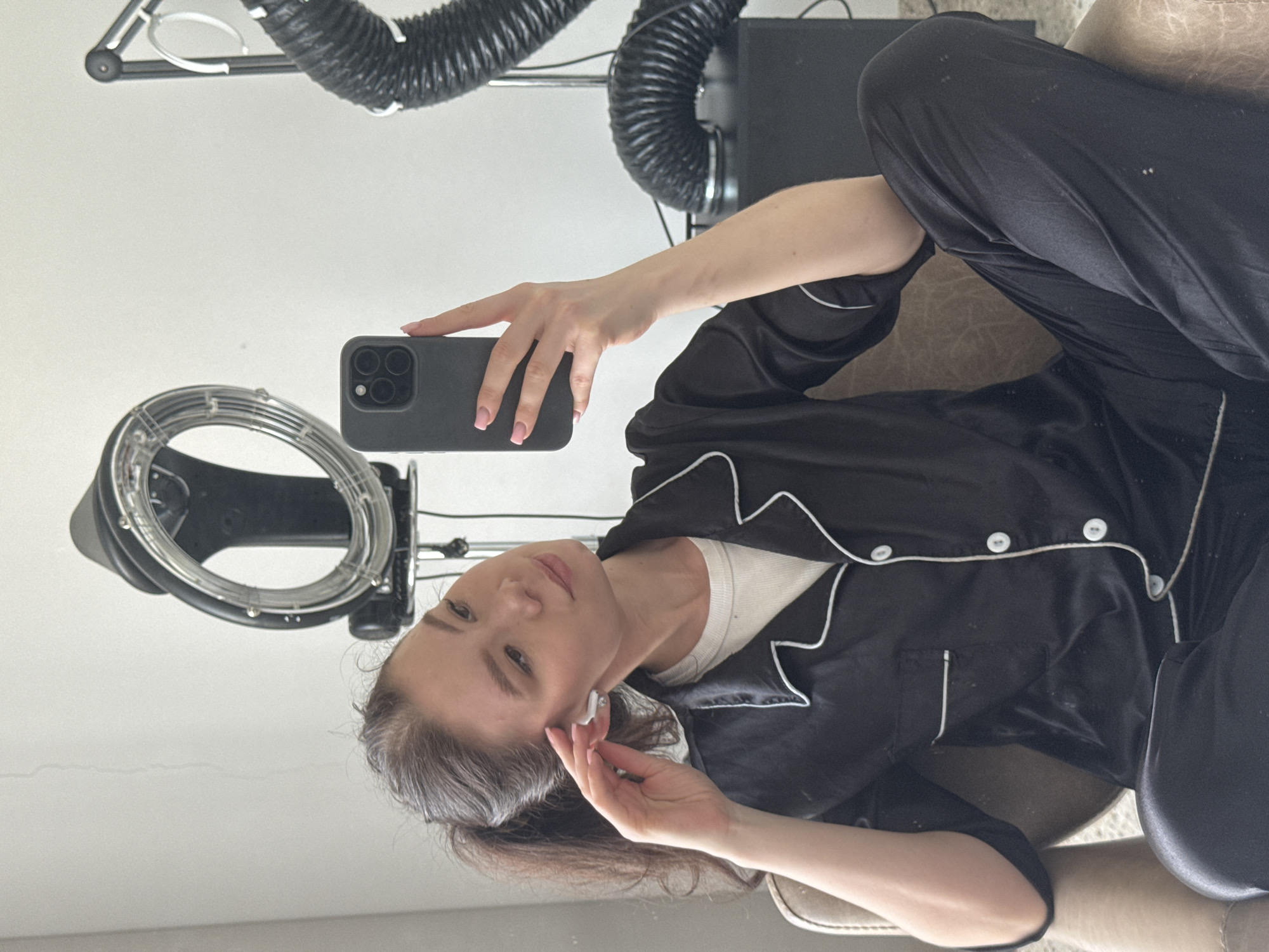 A woman in a black pajama set and white t-shirt sits in a chair, holding a black smartphone. She is holding a white earbud in her other hand and looking at the camera, taking a selfie. Behind her is a hair dryer, a black box, and some tubing. The location is Unknown, Unknown.