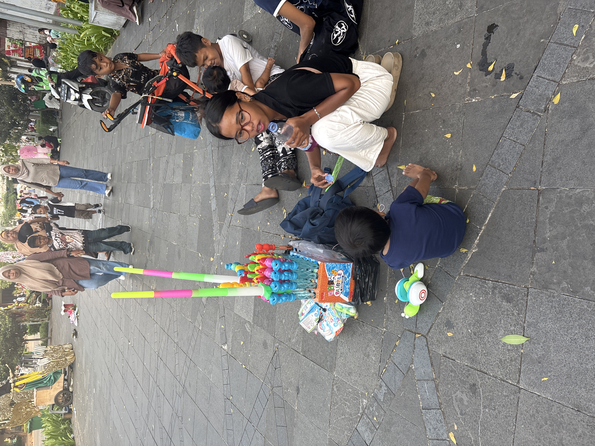 In the city of West Jakarta, Indonesia, a street scene unfolds with several individuals and a variety of objects. In the foreground, a young child wearing a dark blue t-shirt sits on the pavement, facing away from the camera. To the left of the child, a green and white toy with red circular details lies on the ground. A cart filled with colorful bubble wands and boxes, including one labeled "BUBBLE STICK," is positioned in the middle ground. A young woman wearing glasses, a black t-shirt, and white pants sits on the pavement, holding a clear plastic bottle and its cap. Another child, wearing a white t-shirt, is seated next to her, holding a baby. Behind them, two adults are walking away from the camera, one wearing a brown headscarf and a brown tunic with blue jeans, and the other wearing a light brown headscarf and a patterned outfit. In the background, a man on a motorcycle with a blue helmet is visible, along with other people and a stroller. The scene takes place on a paved walkway with scattered yellow leaves.