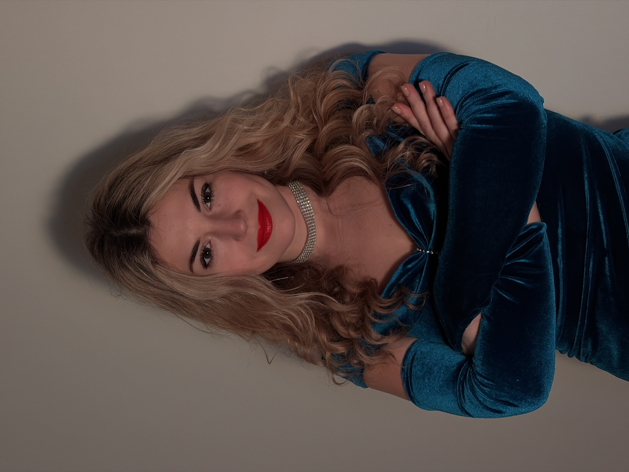 A woman with fair skin, long blonde curly hair, and red lipstick is reclining against a neutral, light gray wall. She is wearing a teal velvet dress with long sleeves and a bow at the neckline, along with a sparkling choker necklace. Her arms are crossed. It's impossible to determine the location of the photograph as being in the city of Budva, Montenegro as there are no background clues to that effect.