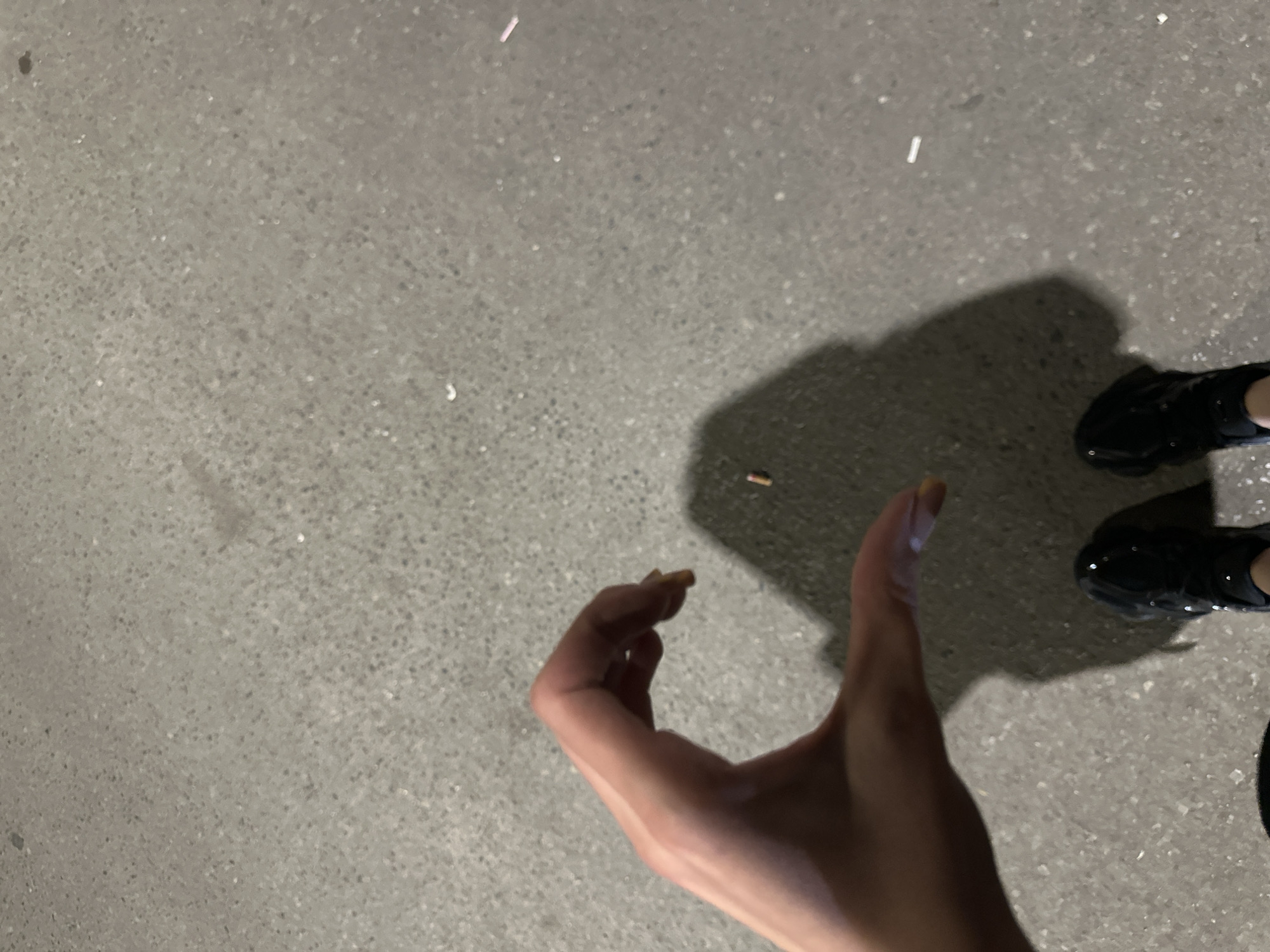 A person's left hand forms the shape of a heart, with the thumb and index finger extended. The hand casts a shadow on a grey asphalt surface. Also on the asphalt are two cigarette butts. The feet of the person, wearing black sneakers, are visible at the bottom of the frame. The scene is outdoors, possibly at night due to the shadow, with no other discernible environmental features.