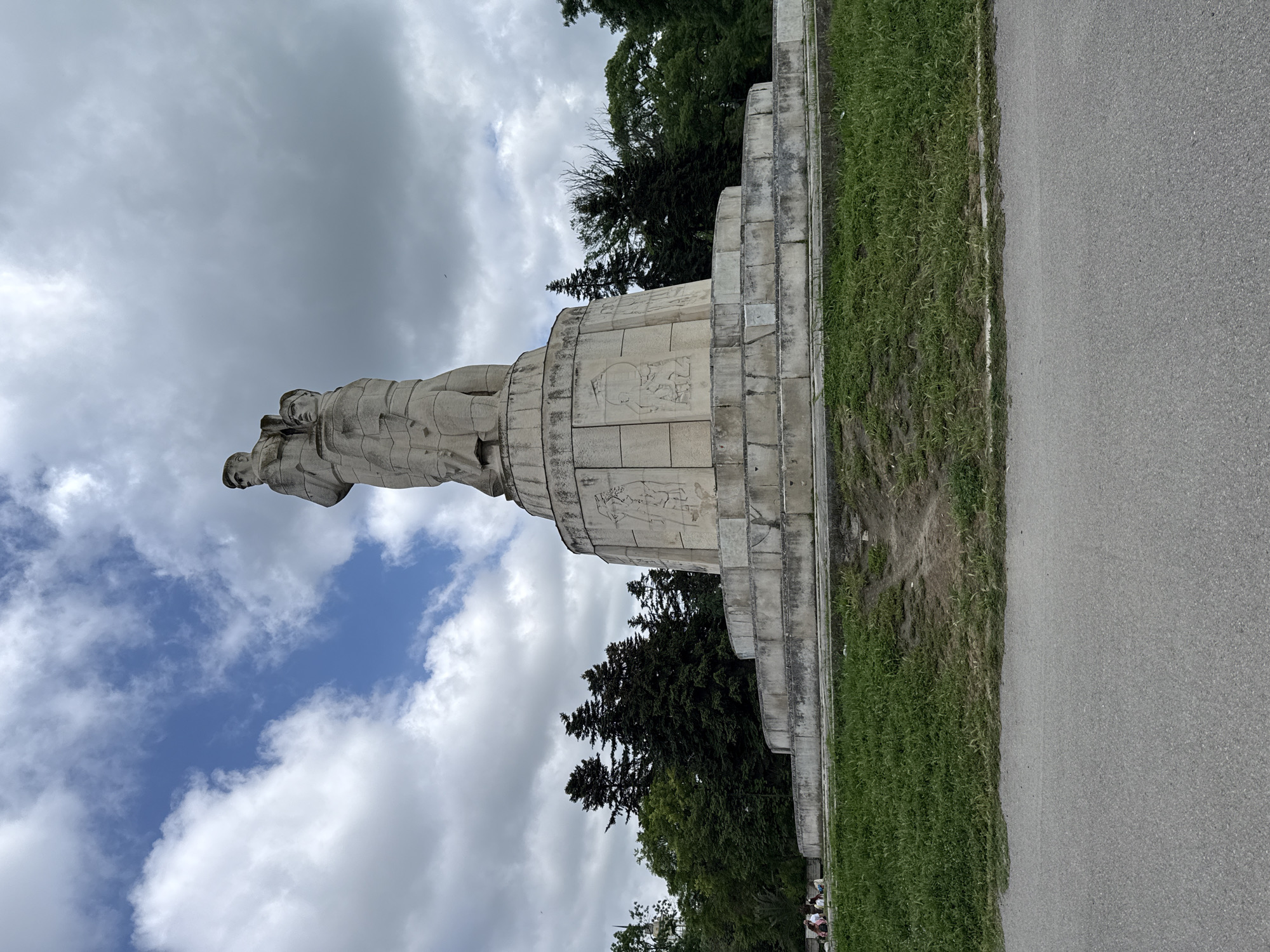 Here's a description of the image:
The image shows a large stone monument in what appears to be a park-like setting. The monument consists of a raised, tiered stone platform upon which two figures stand. The figures appear to be male, and they are depicted in a somewhat stylized, heroic style. The base of the monument is adorned with bas-relief carvings. A paved path runs adjacent to the monument. The sky is overcast with clouds, but some patches of blue are visible. Trees are visible in the background. There are no people shown in the foreground but some indistinct groups are shown in the bottom right. The location is listed as Unknown, Unknown.