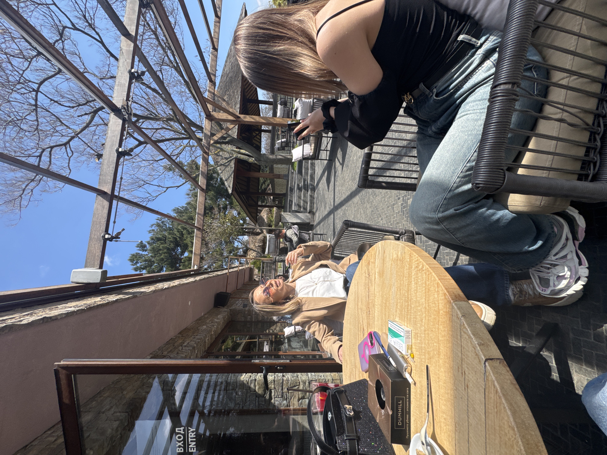 A sunlit outdoor scene depicts two women at a café in Varna, Bulgaria. One woman, wearing a black top and jeans, sits in a wicker chair, angled towards the other. She is holding a phone, possibly taking a picture. The second woman, wearing a tan jacket over a white top, is seated at a round wooden table. Sunglasses rest on her face and she is smiling broadly. On the table, there is a box labeled "DUNHILL", various personal items, and what appears to be medication. Behind them, a stone building is visible through a large window, with an "ENTRY" sign displayed. The café is shaded by a wooden pergola structure. Other patrons and building structures are visible in the background.