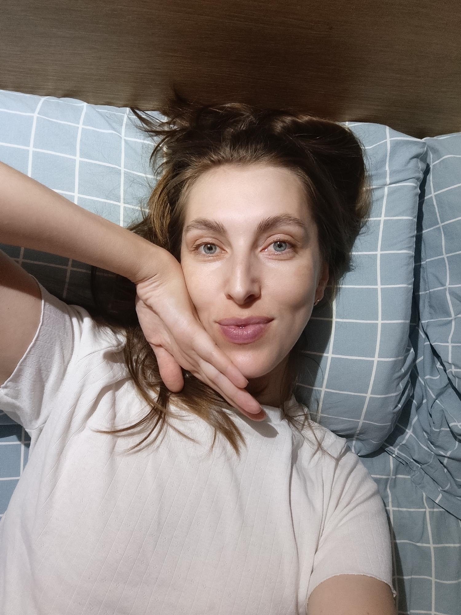 The image shows a young woman lying on a bed, looking at the camera. She is wearing a white t-shirt and has her hair spread out on the pillow. Her left arm is raised, with her hand resting on her cheek. The pillow has a blue and white checkered pattern. The background appears to be a wooden headboard. The lighting suggests an indoor setting, possibly a bedroom. There is no indication of the location being in Chaikovsky, Russia, or any other specific city.