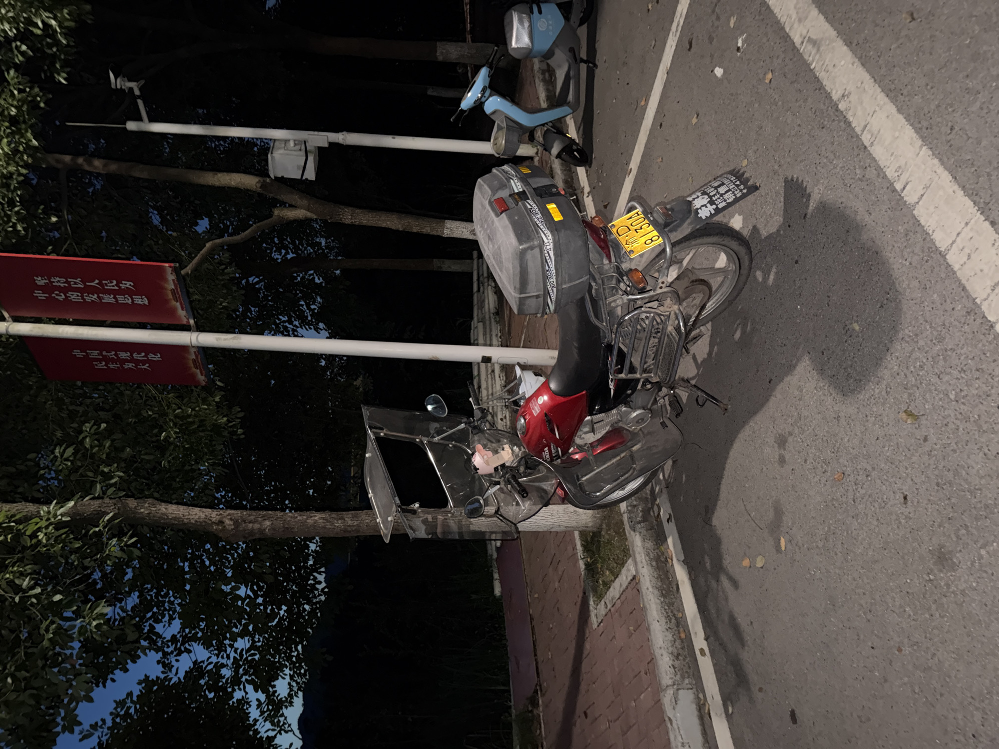A red Honda motorcycle, fitted with a large transparent windshield and a black storage box, is parked on an asphalt surface next to a sidewalk made of red bricks. The motorcycle's license plate reads "渝D 8130A". To the right of the motorcycle is a blue electric scooter. In the background, there are trees and a lamppost with a small white box attached. Red banners with Chinese characters hang from poles to the left of the trees. The scene appears to be outdoors in a city location at dusk or night. The area is likely the city of Jiangjin, China, given the format of the license plate.