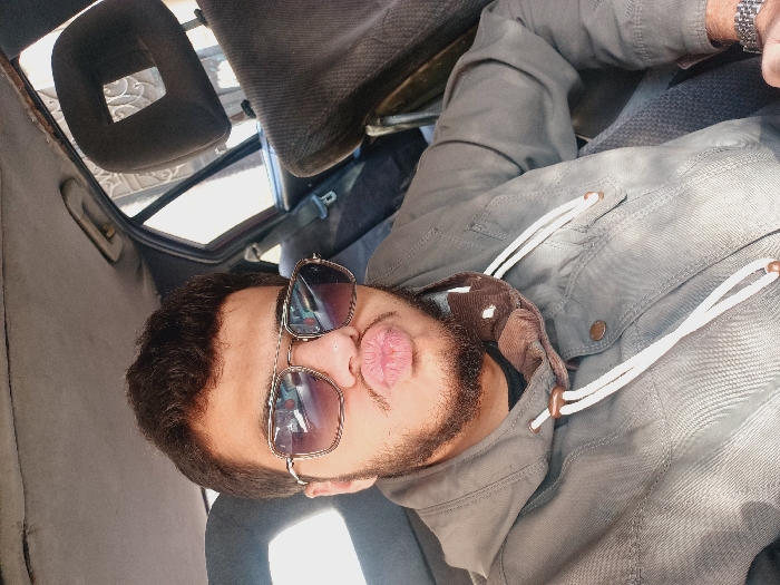 A man sits in a car wearing sunglasses and a grey jacket with white drawstrings. He's looking at the camera with pursed lips. The interior of the car is visible, including the seats, headrests, and the car's ceiling. Light filters in through the window. A wristwatch is visible on a hand in the corner of the image.