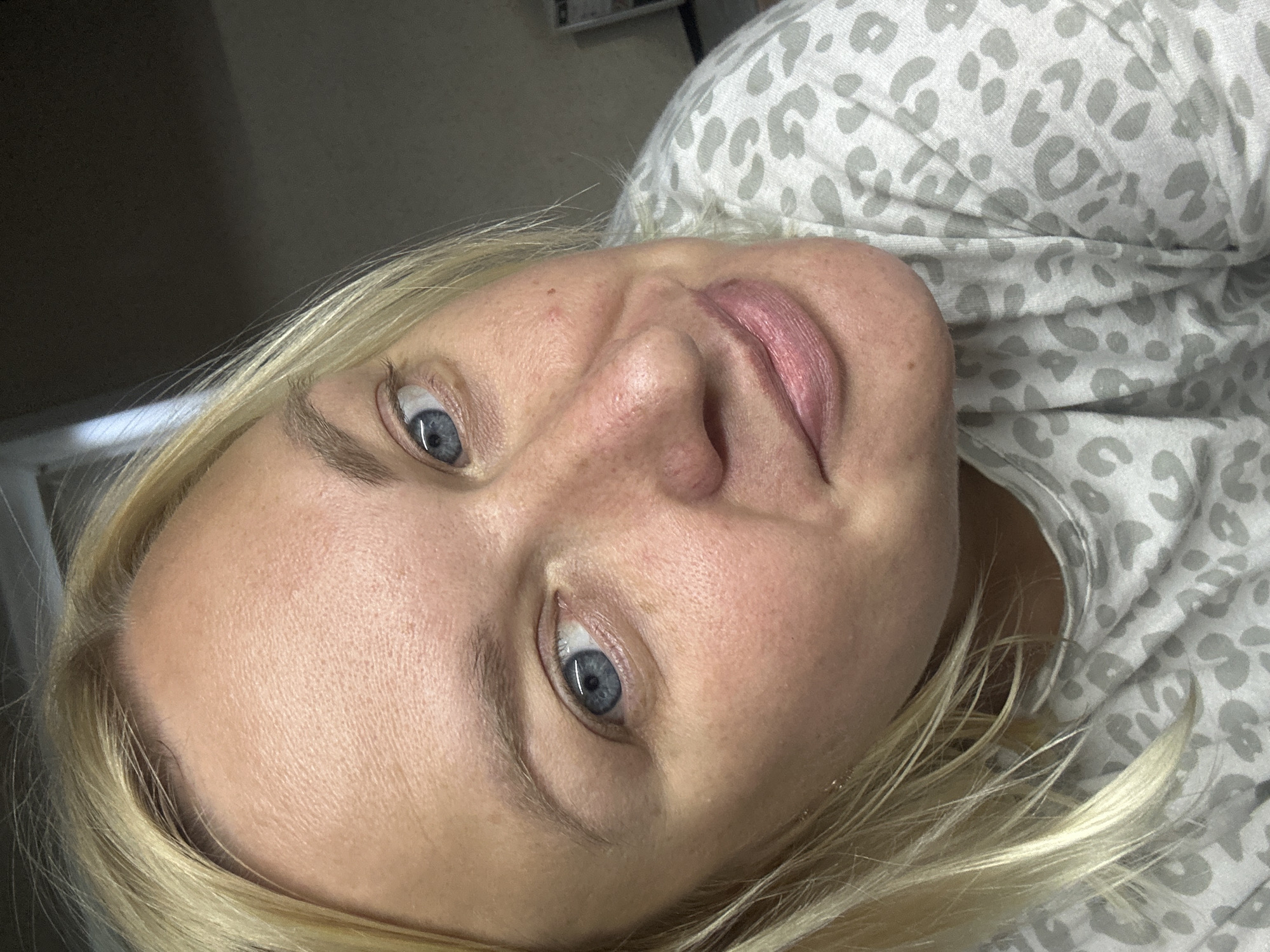 A close-up, high-angle, indoor shot shows a fair-skinned woman with blonde hair and blue eyes. She is wearing a light-colored shirt with a gray leopard print pattern. The background is dark and out of focus, with a hint of a window on the upper left. The image appears to have been taken in Chaikovsky, Russia, within a residential or personal space.